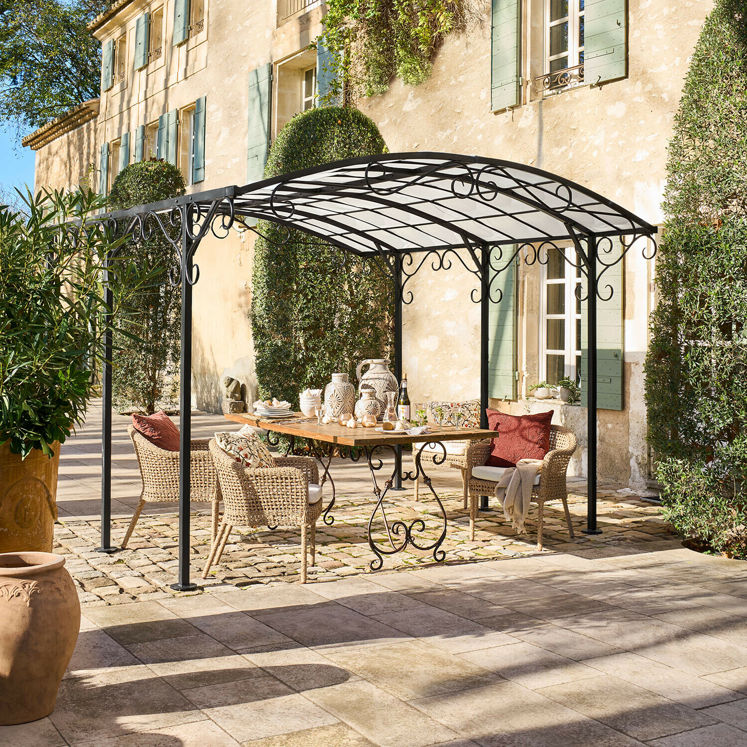 Pergola Delbridge Zwarte metalen tuinprieel met sierlijke krullen boven houten tafel en rotan stoelen op terras