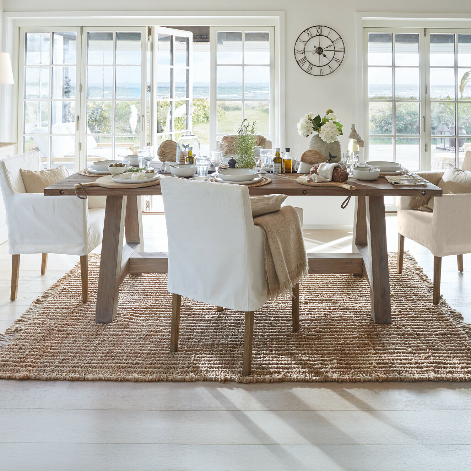 Eetkamer met houten tafel en witte stoelen op natuurlijke vloerkleed met uitzicht op tuin en zee