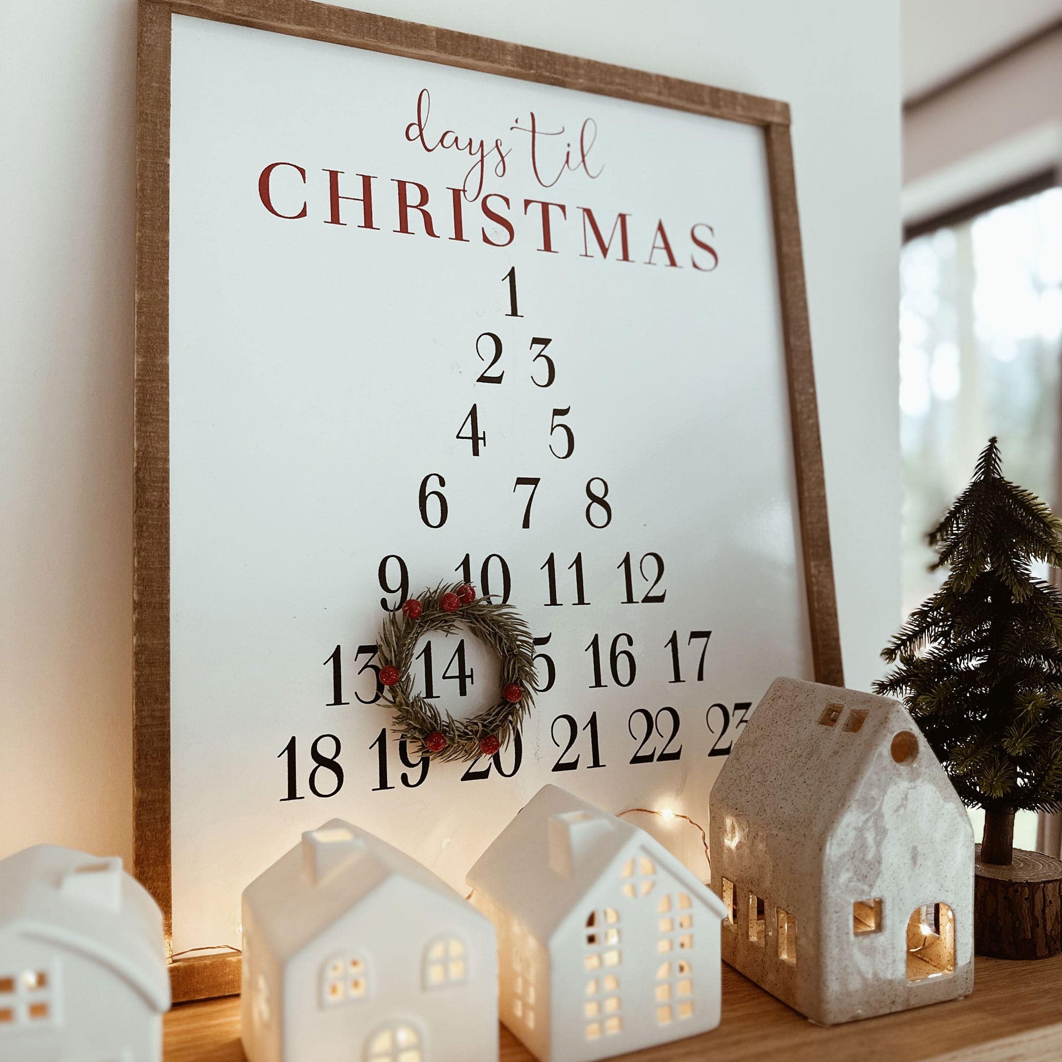Houten lijst met adventskalender en decoratieve krans, keramische huisjes en kerstboom op tafel