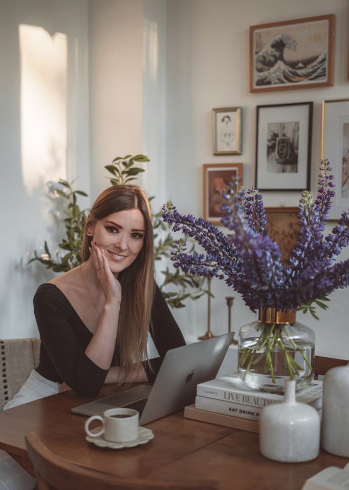 Vrouw aan houten tafel met laptop en glazen vaas met paarse bloemen in lichte kamer
