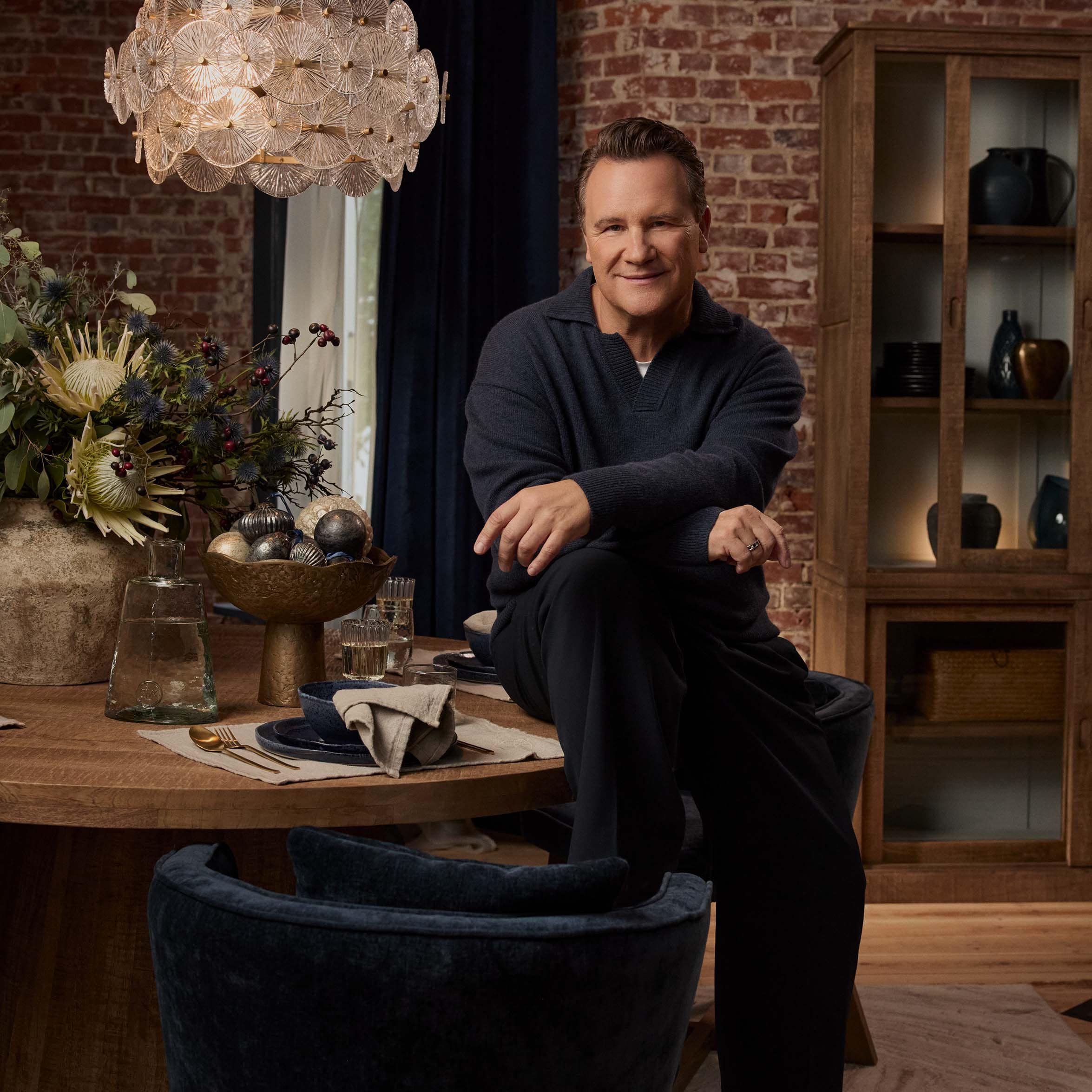 Man in donkerblauwe trui zittend aan houten tafel met decoraties en lamp van transparante glazen schijven