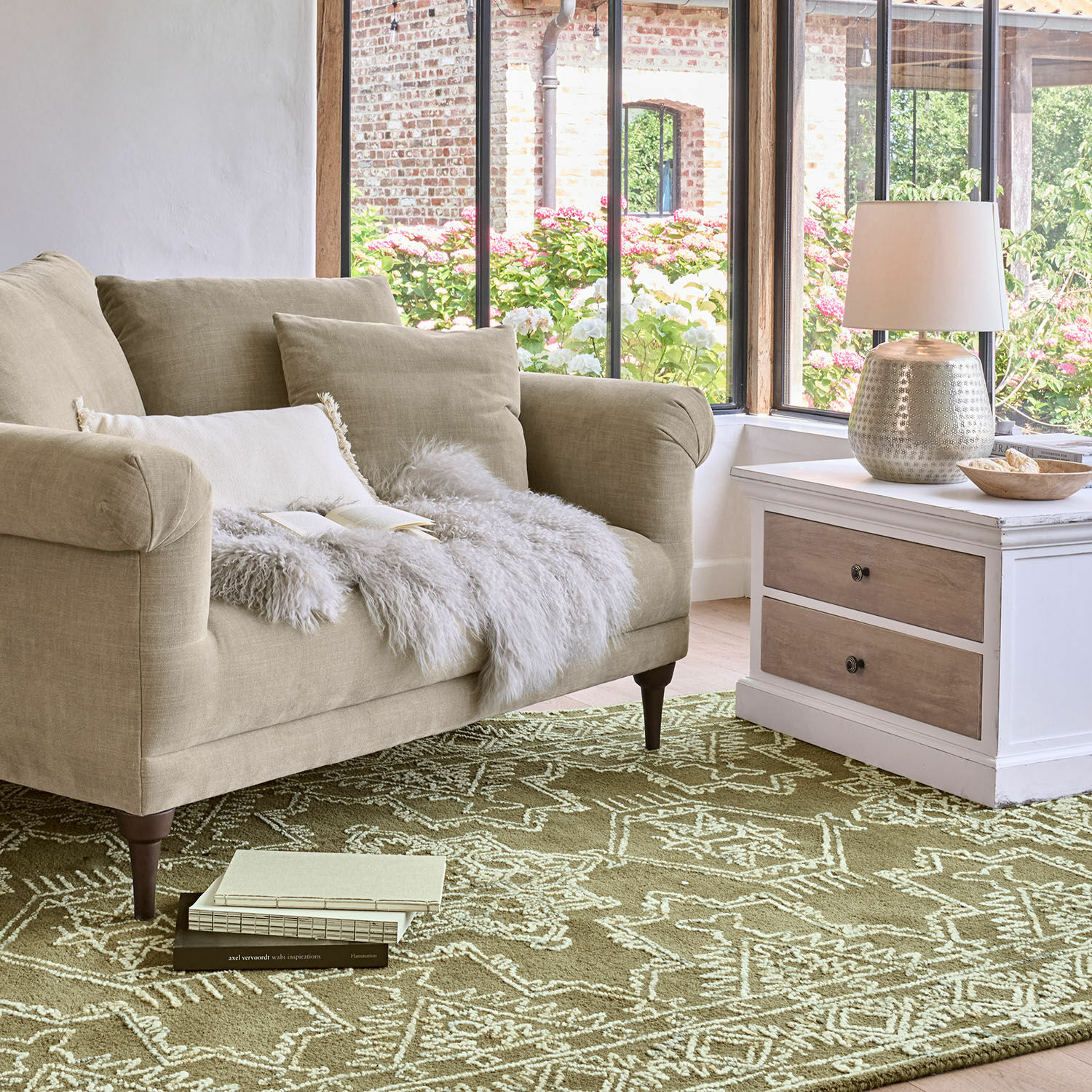 Beige stoffen bank met kussens en een bontdeken op een groen vloerkleed met geometrisch patroon