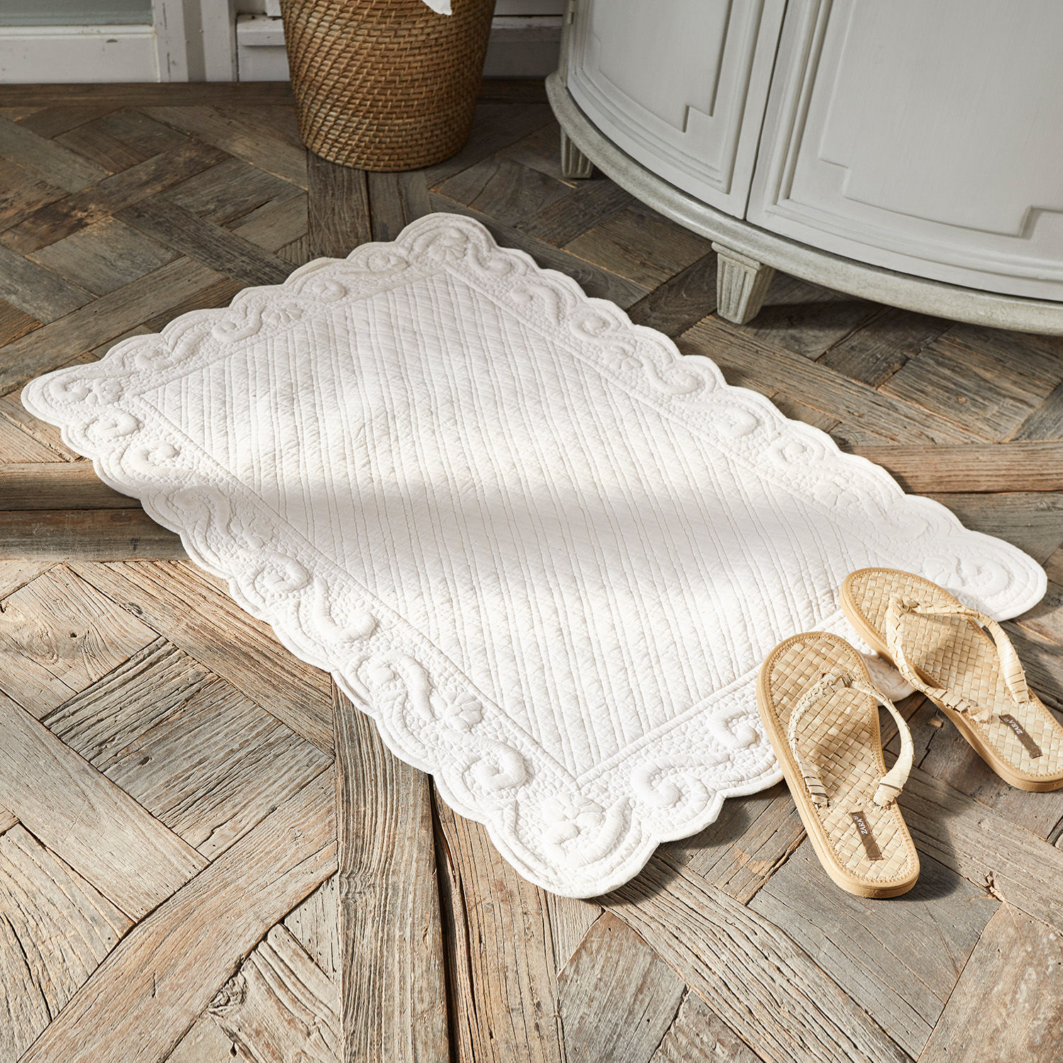 Badmat Svikola Witte decoratieve katoenen mat met doorgestikt patroon op houten vloer naast gevlochten slippers