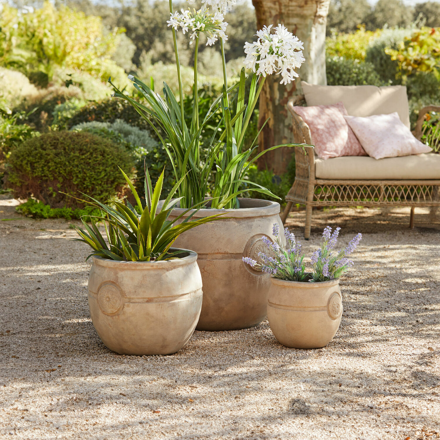 Drie beige terracotta bloempotten met planten op grindpatio in tuin met rotan bank