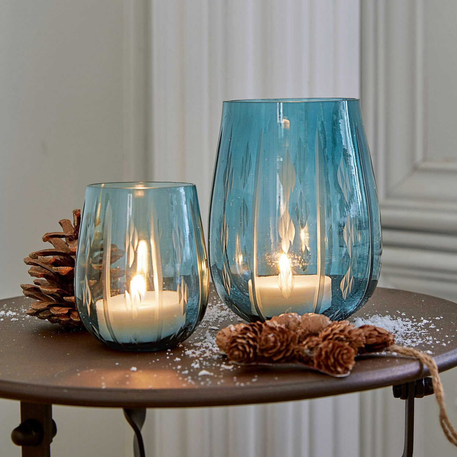 Windlicht set van 2 Danisar Twee blauwe glazen windlichten met patroon, witte kaarsen en dennenappeldecoratie op tafel