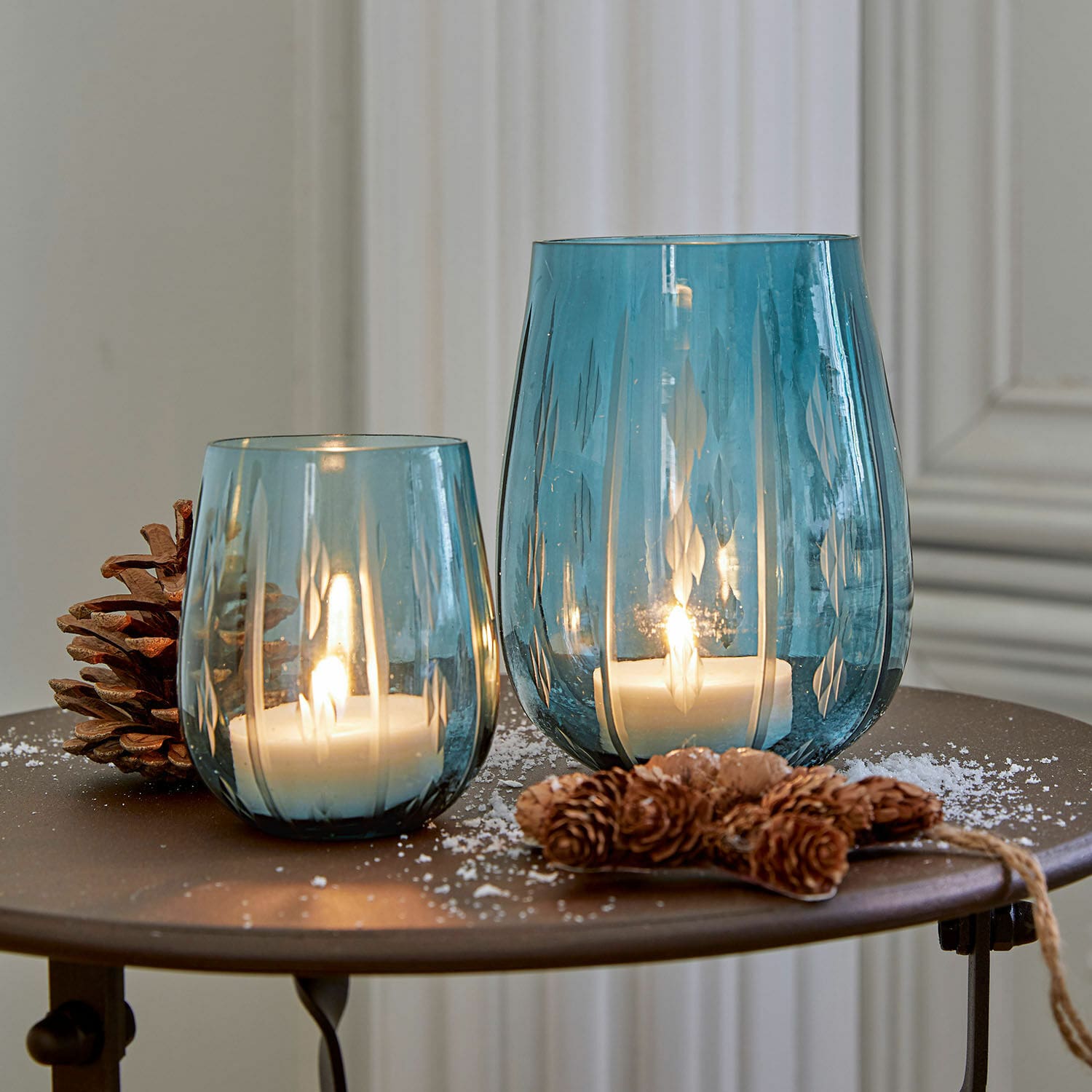 Windlicht set van 2 Danisar Twee blauwe glazen windlichten met patroon, witte kaarsen en dennenappeldecoratie op tafel