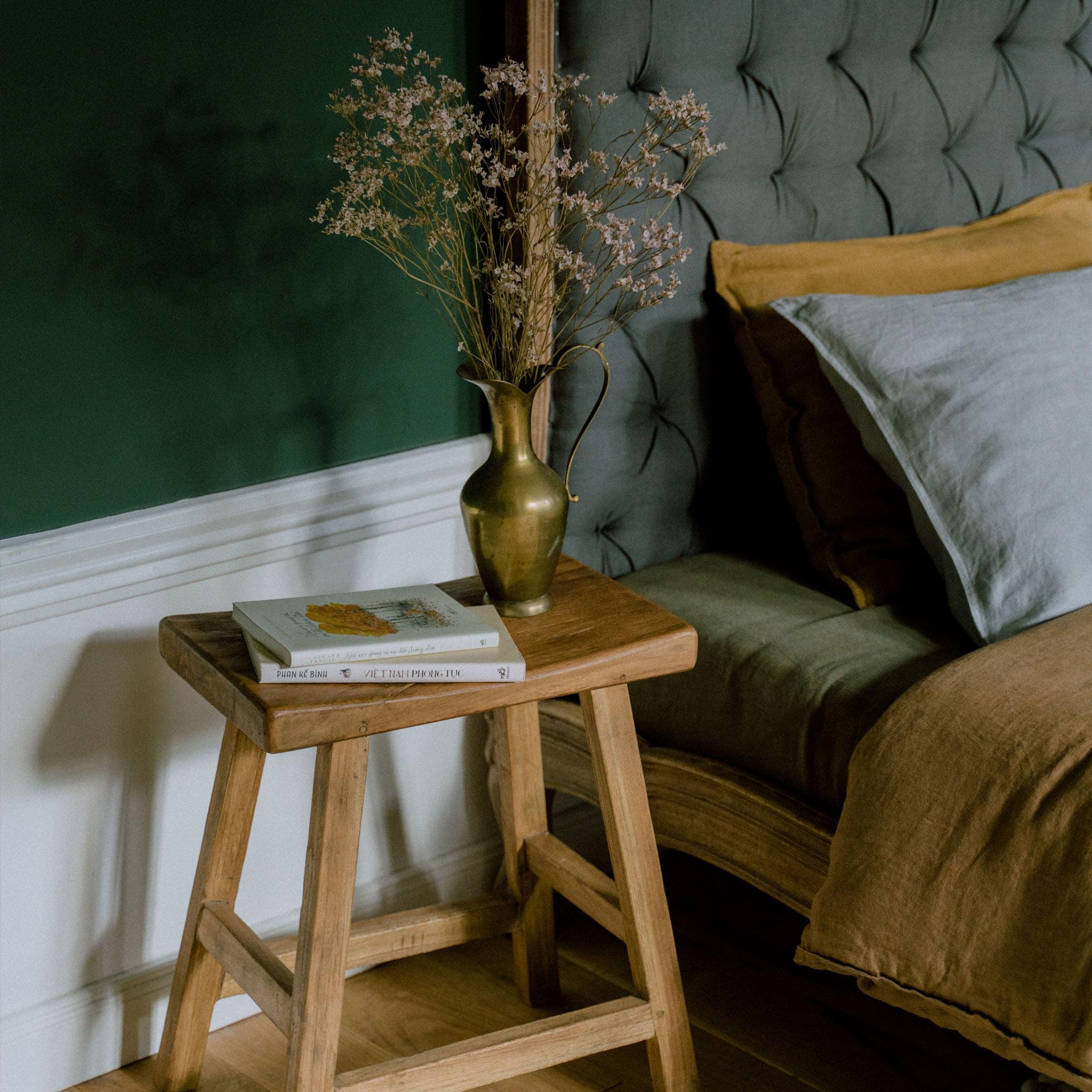 Houten kruk met messing vaas en gedroogde bloemen naast een bed met grijze bekleding en bruin beddengoed
