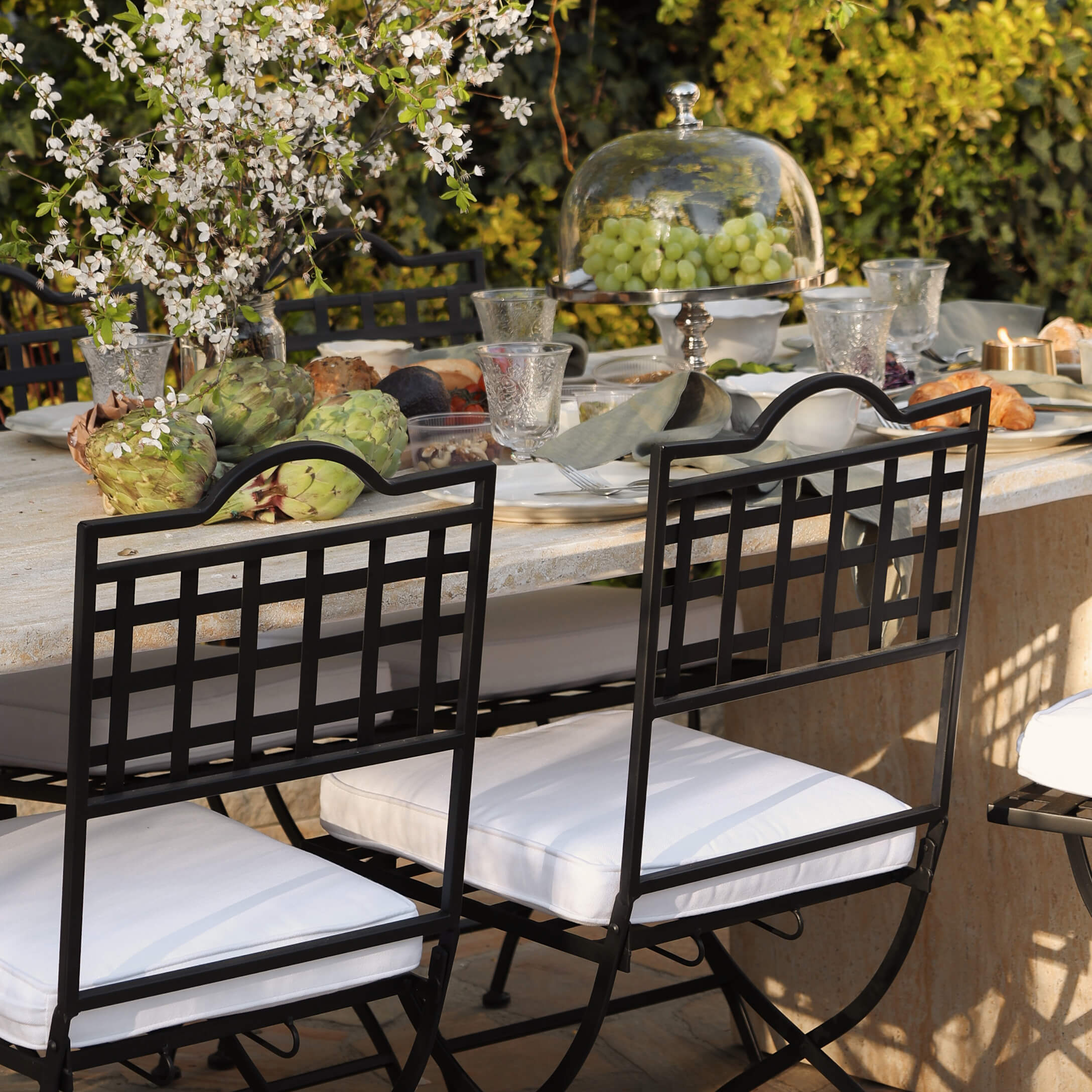 Zwarte metalen stoelen met witte kussens bij stenen tafel met decoratie en eten buiten