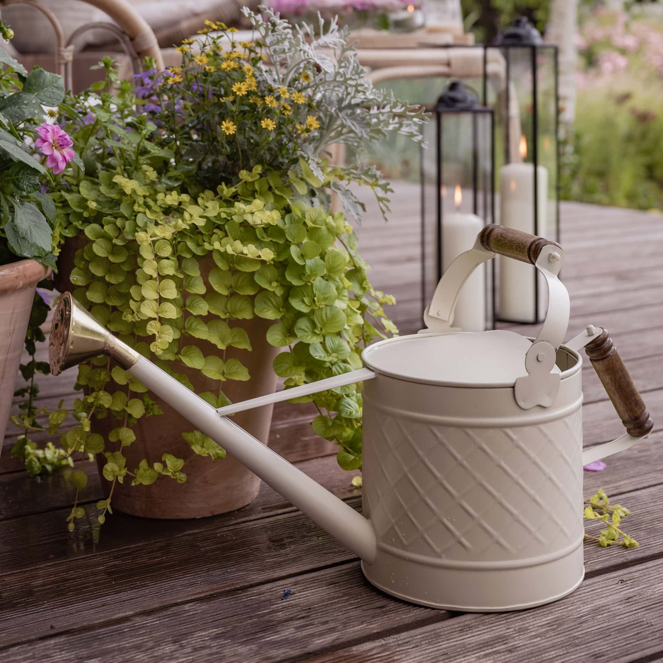Crèmekleurige metalen gieter met houten handvatten op houten terras naast planten en kaarsen