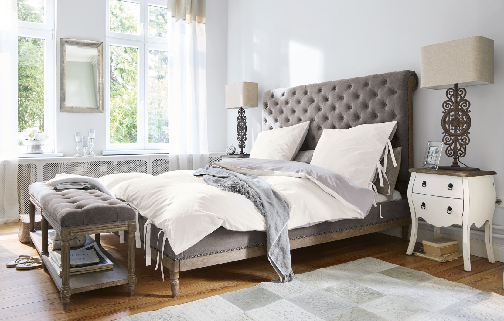 Klassieke slaapkamer met grijs gestoffeerd bed en wit beddengoed in lichte ruimte
