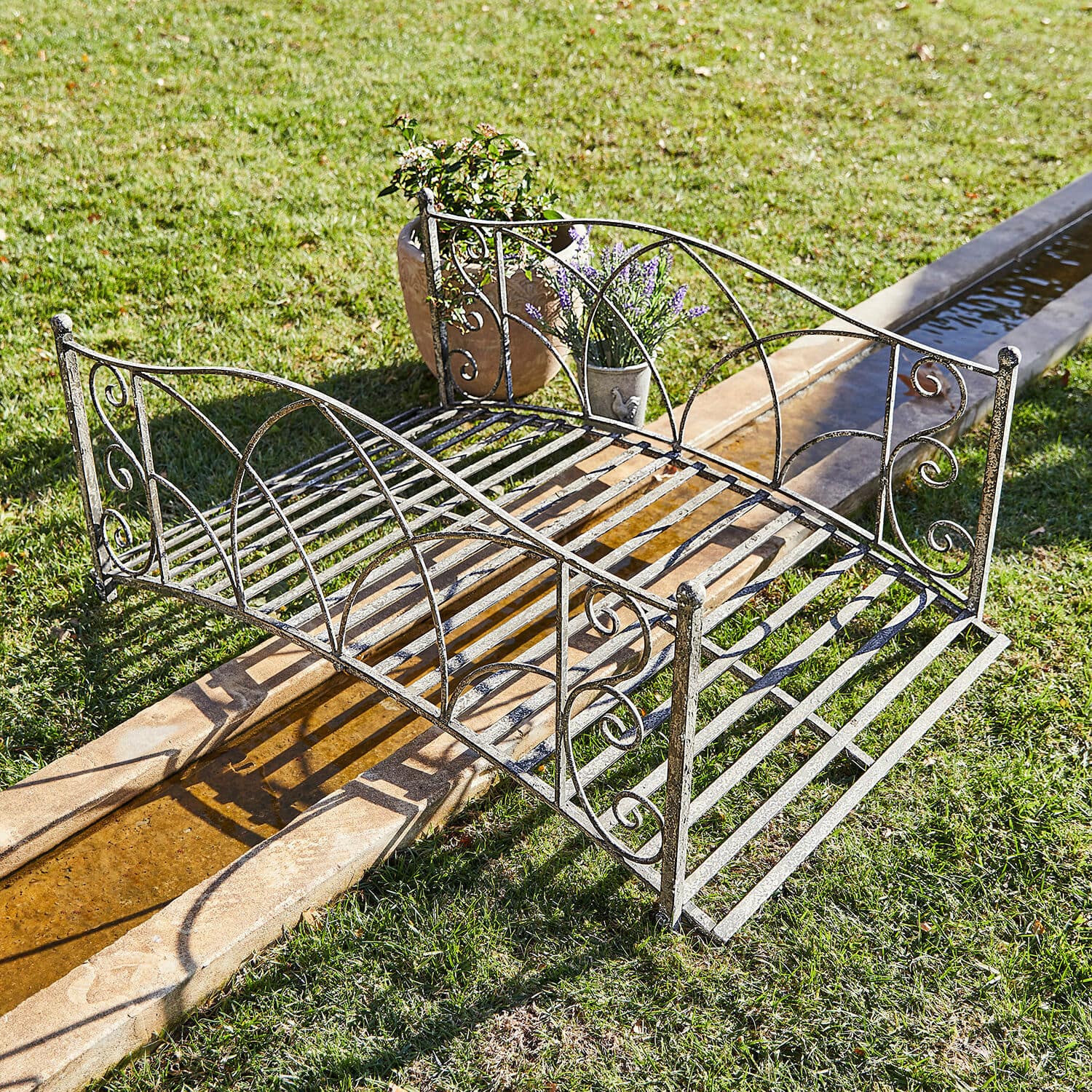 Decoratieve brug Escàs Sierlijke metalen tuinbrug in verouderd zilver over een waterloop op het gras met bloempotten