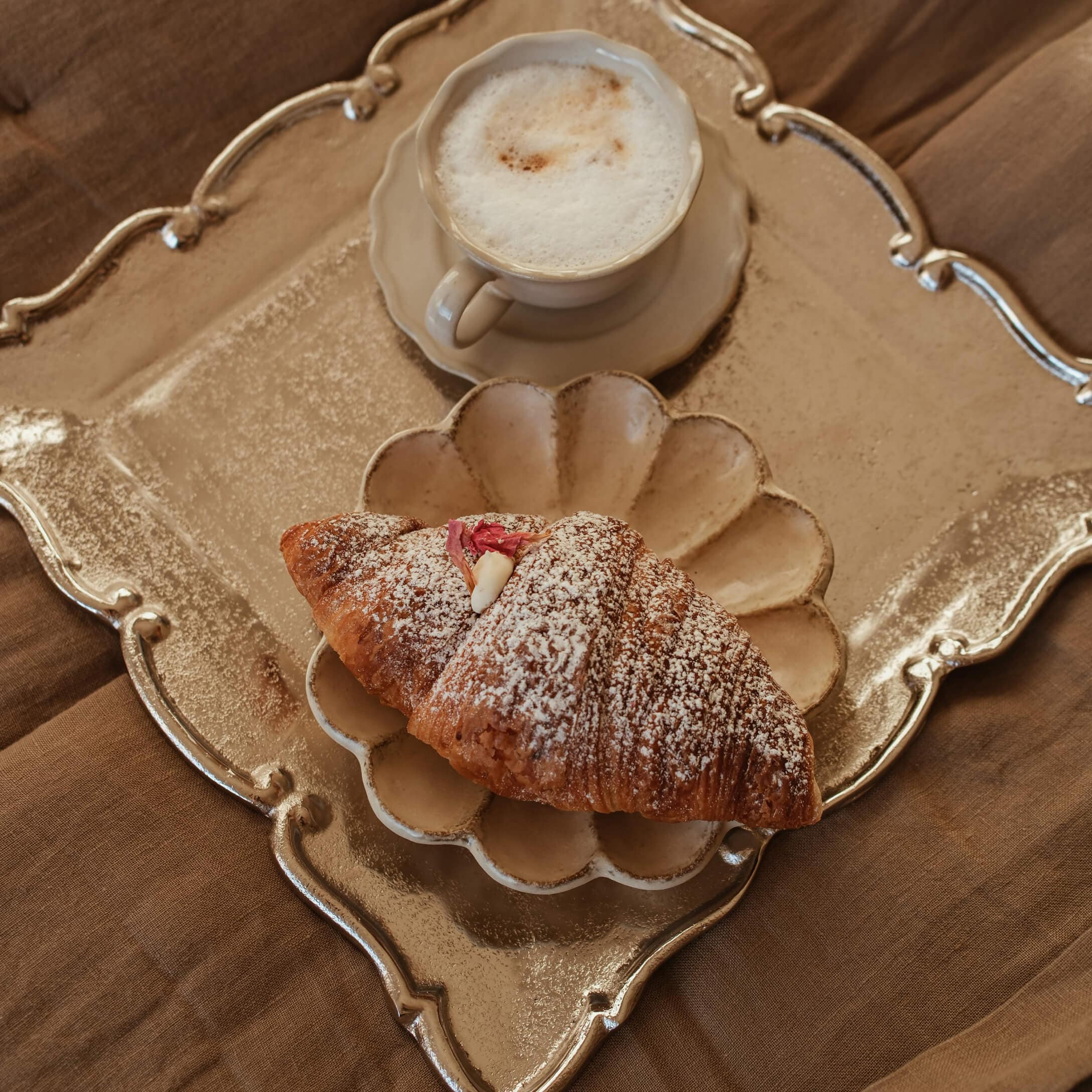 Kopje romige cappuccino en croissant met poedersuiker op beige bord en gouden dienblad