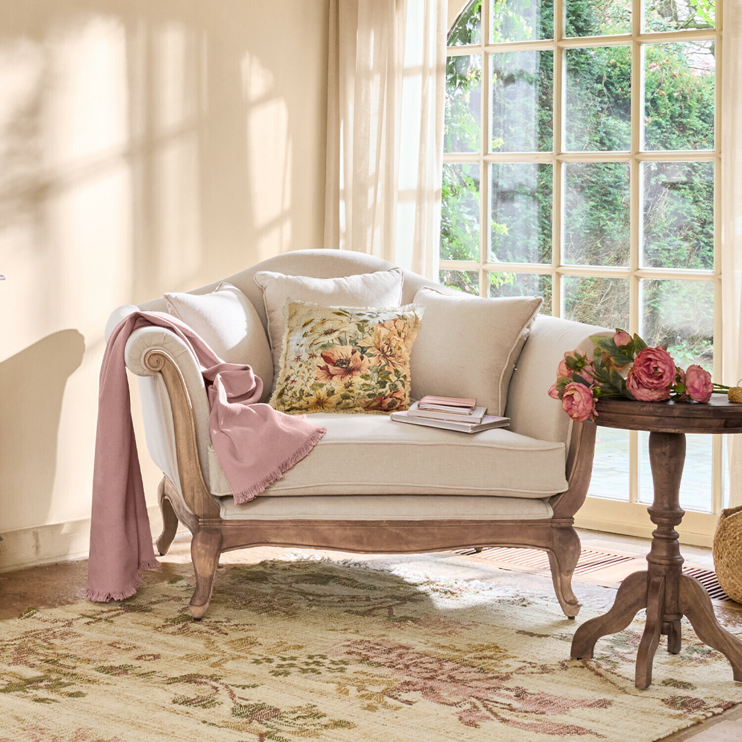 Fauteuil Sumerset Beige bank met houten frame, bloemenkussen en roze deken in lichte woonkamer met groot raam
