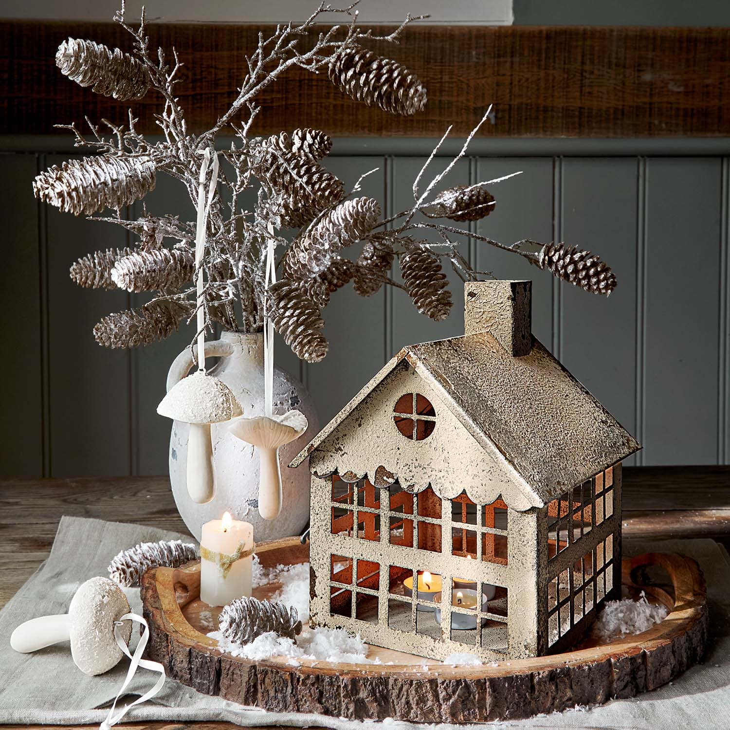 Decoratieve metalen lantaarn in huisvorm beige met dennenappeltakken en kaars op houten dienblad