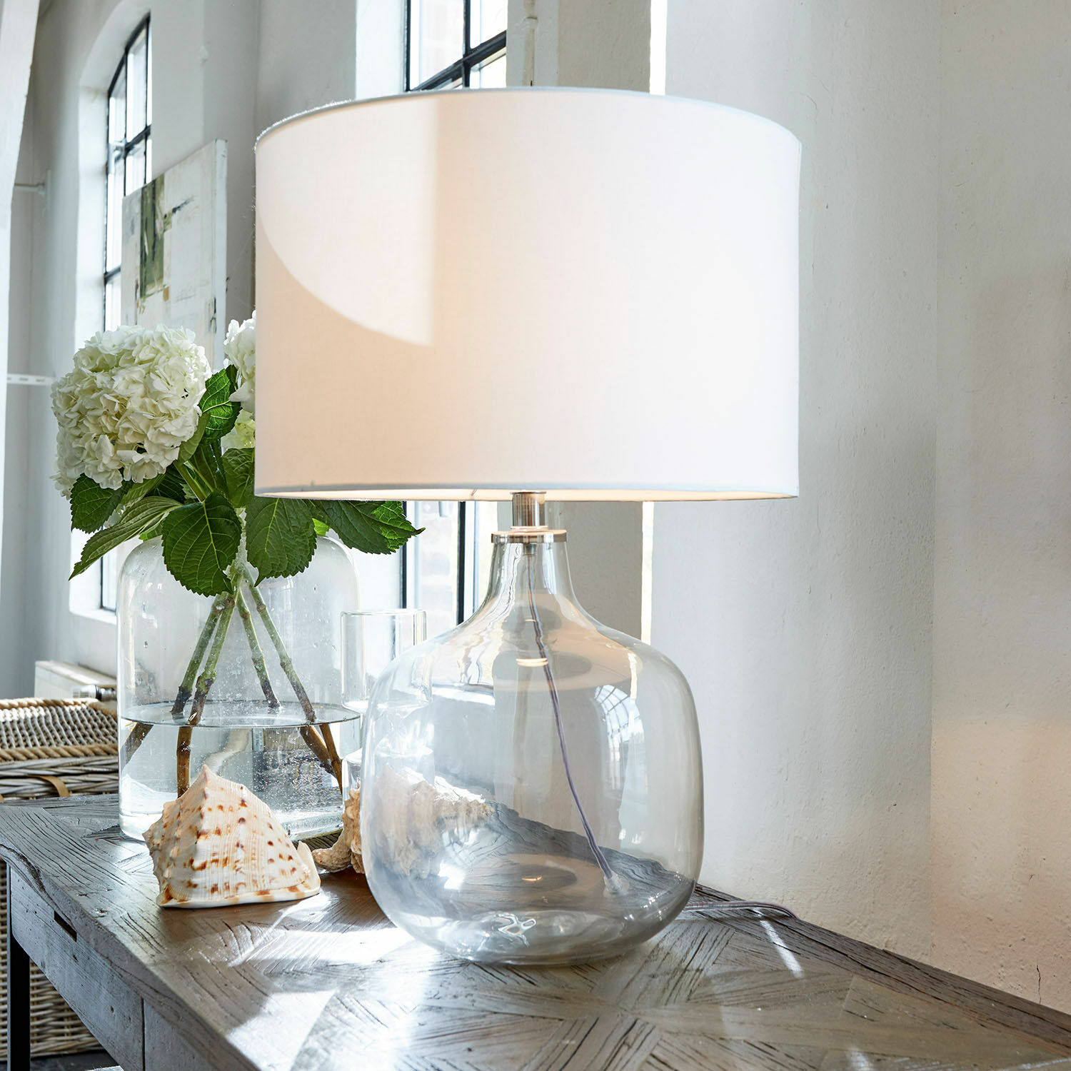 Tafellamp van helder glas met witte kap op houten tafel met schelpen als decoratie