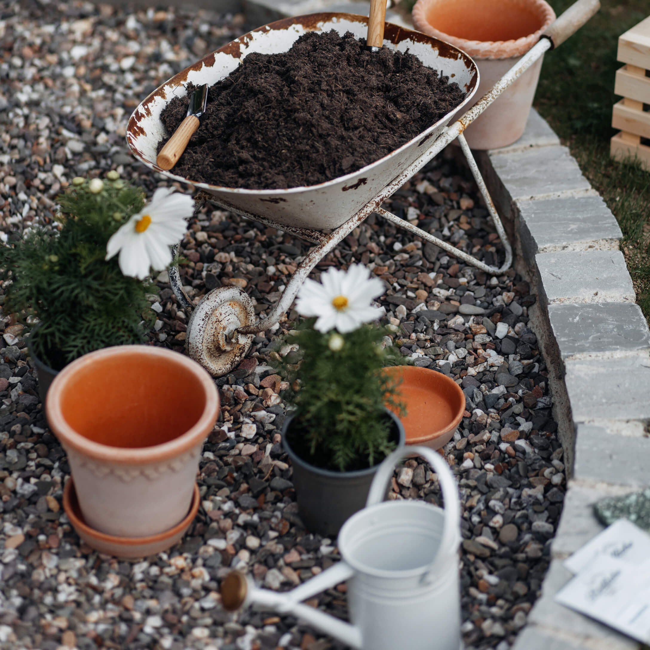 Oude witte kruiwagen met aarde op grind naast bloempotten en gieter in de tuin