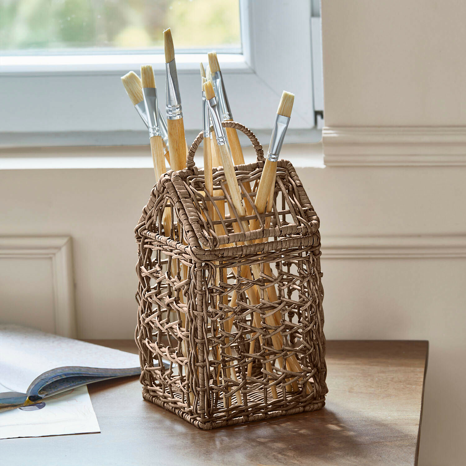 Bruine gevlochten opbergmand in huisvorm met verfkwasten op houten tafel