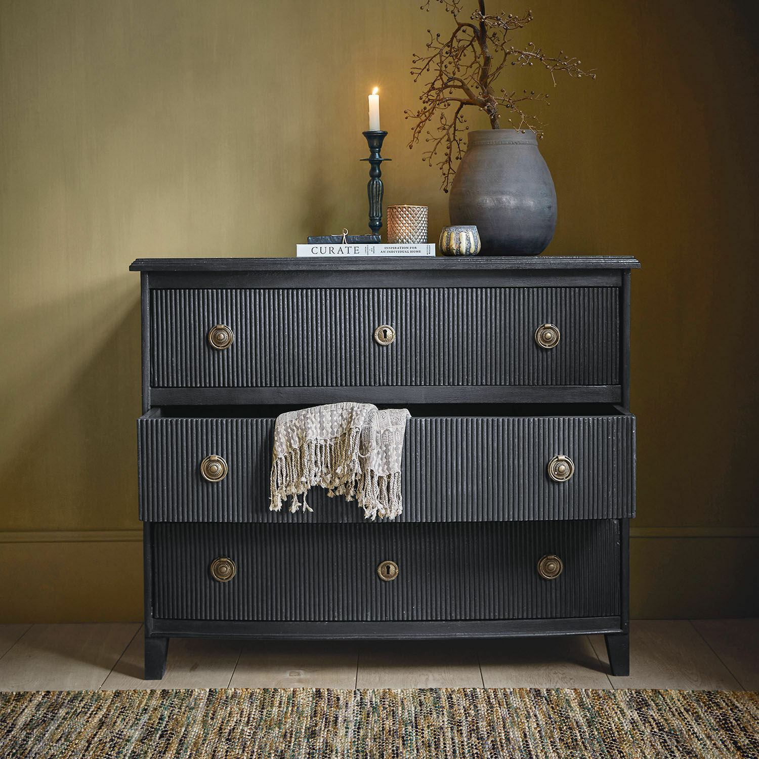 Ladekast Boissiére Zwarte houten ladekast met drie laden en een beige decoratief kleed, vintage stijl