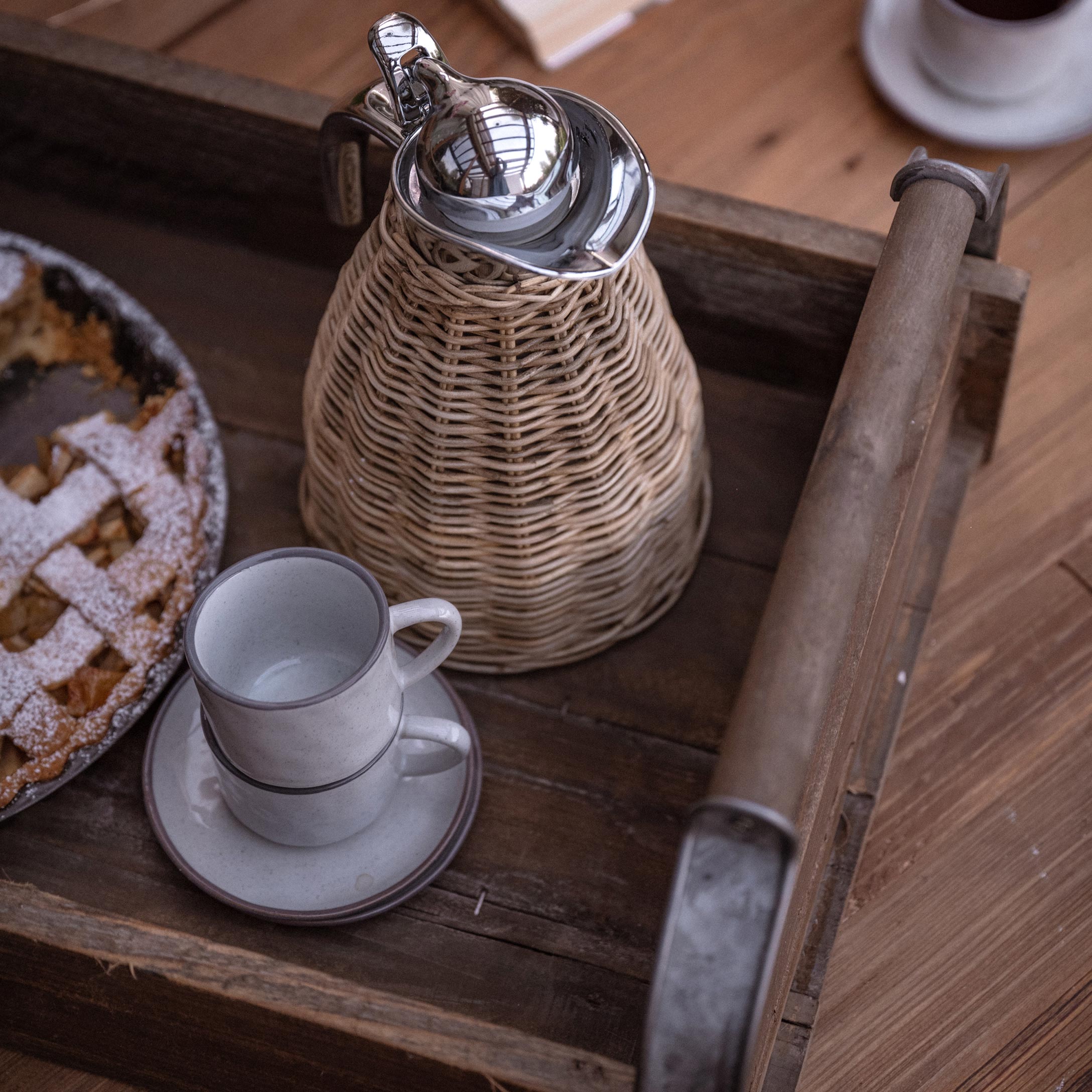Houten dienblad met rieten kan en twee witte kopjes naast taart op houten tafel