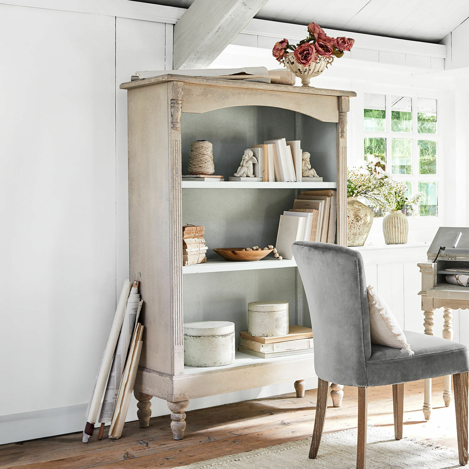 Boekenkast Arterton Beige houten vintage boekenkast met decoratieve boeken en grijze stoel in lichte kamer