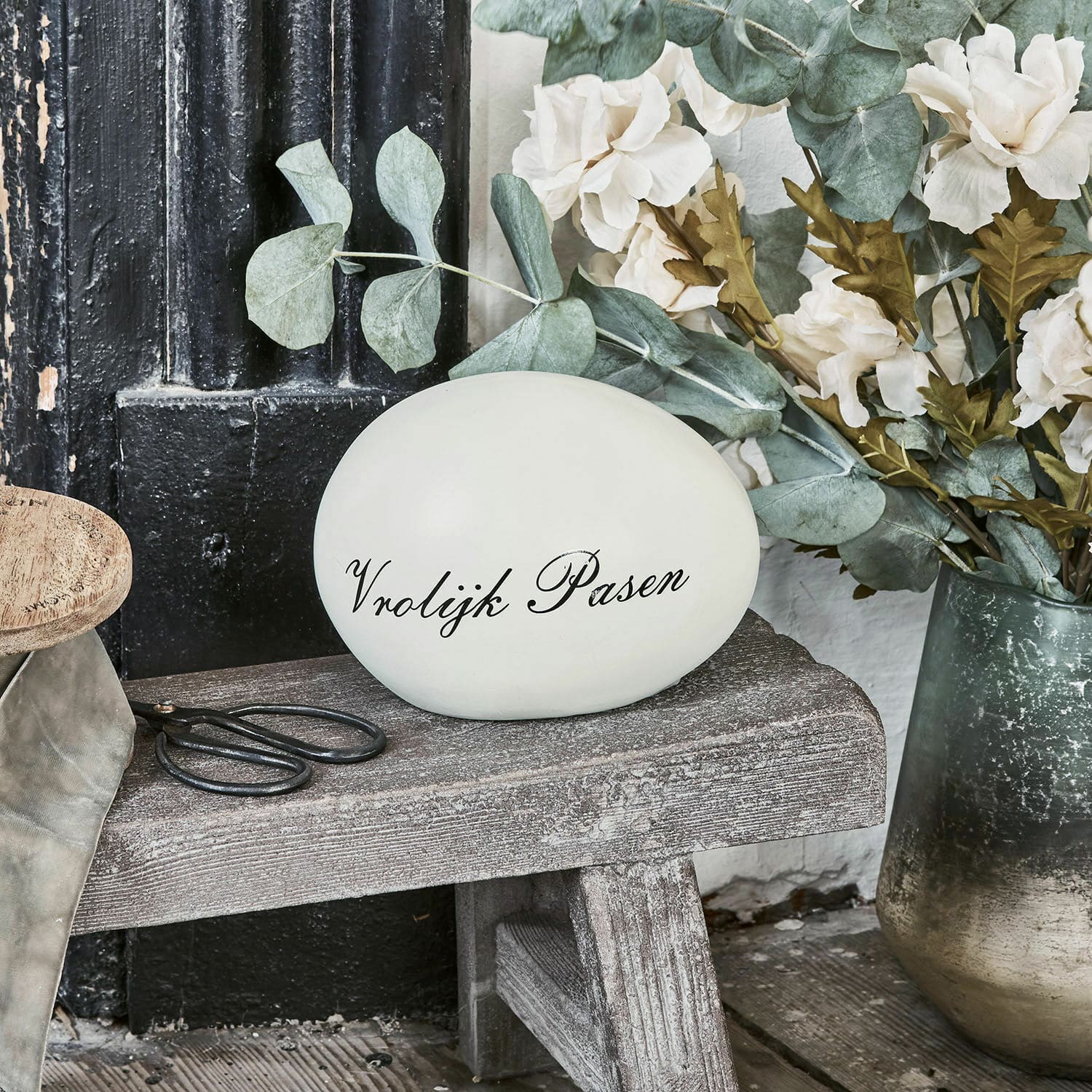 Sierei VROLIJK PASEN Decoratief wit ei met tekst Vrolijk Pasen op houten bankje naast bloemen en schaar