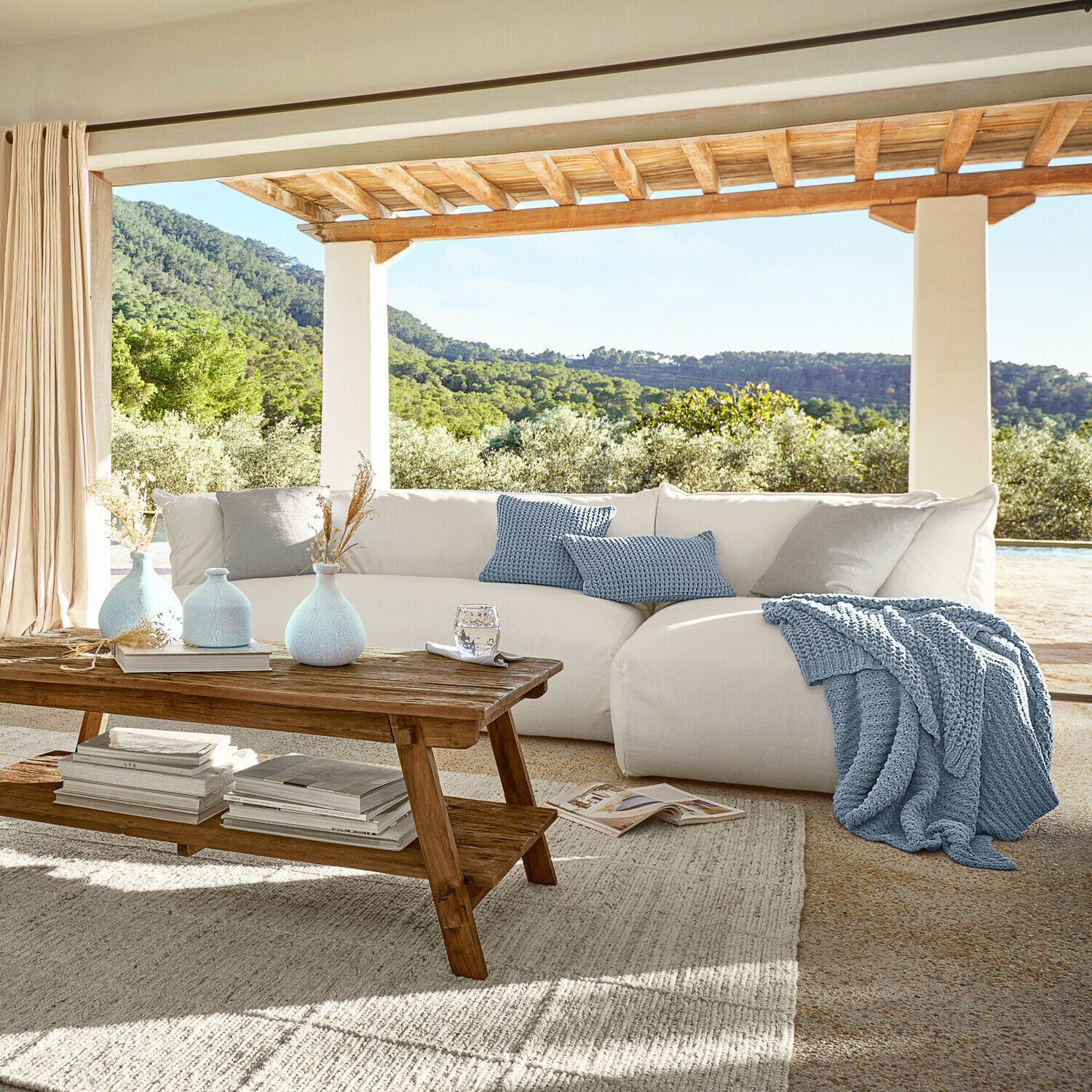 Gezellige witte bank met blauwe gebreide deken en kussens op terras met groen uitzicht