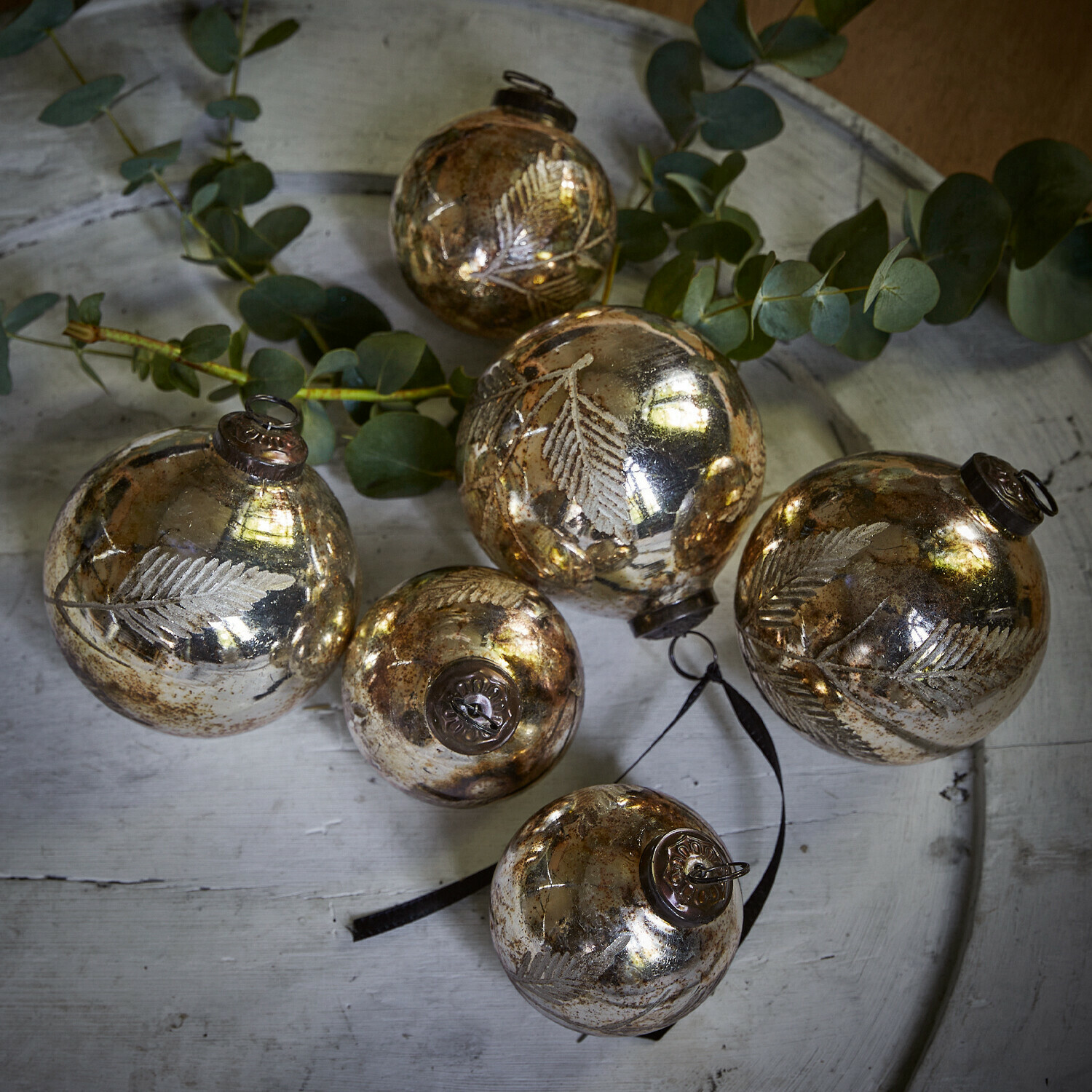 Set van zes gouden glazen kerstballen met bladmotief op een wit dienblad met groene takken