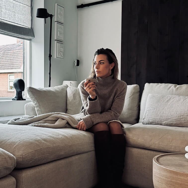 Vrouw in grijze trui zittend op beige bank in moderne woonkamer met groot raam