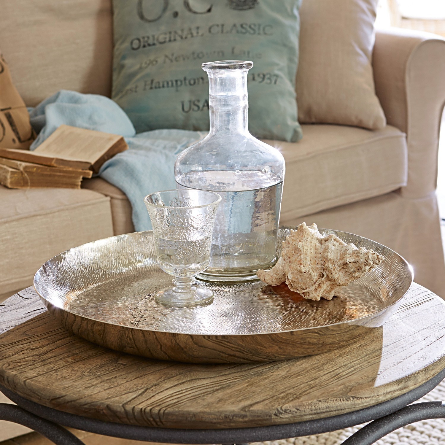 Ronde zilverkleurige metalen dienblad op houten tafel met glazen karaf en schelpdecoratie