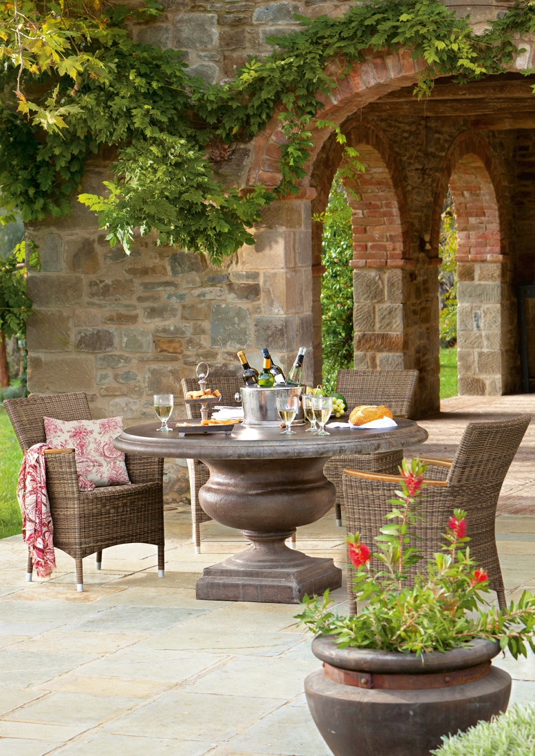 Ronde stenen tuintafel met rieten stoelen op stenen terras met bogen en groenblijvende planten