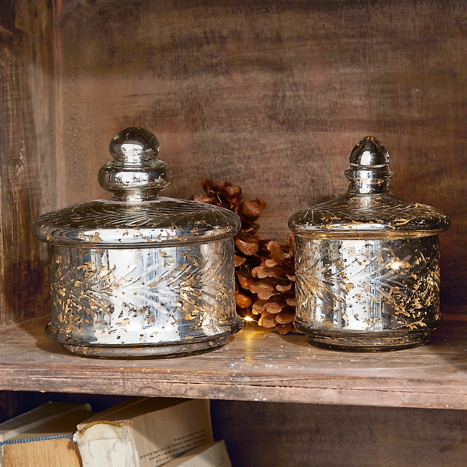Glazen pot set van 2 Elexa Twee glazen potten met deksels en gouden versieringen op houten plank met dennenappels