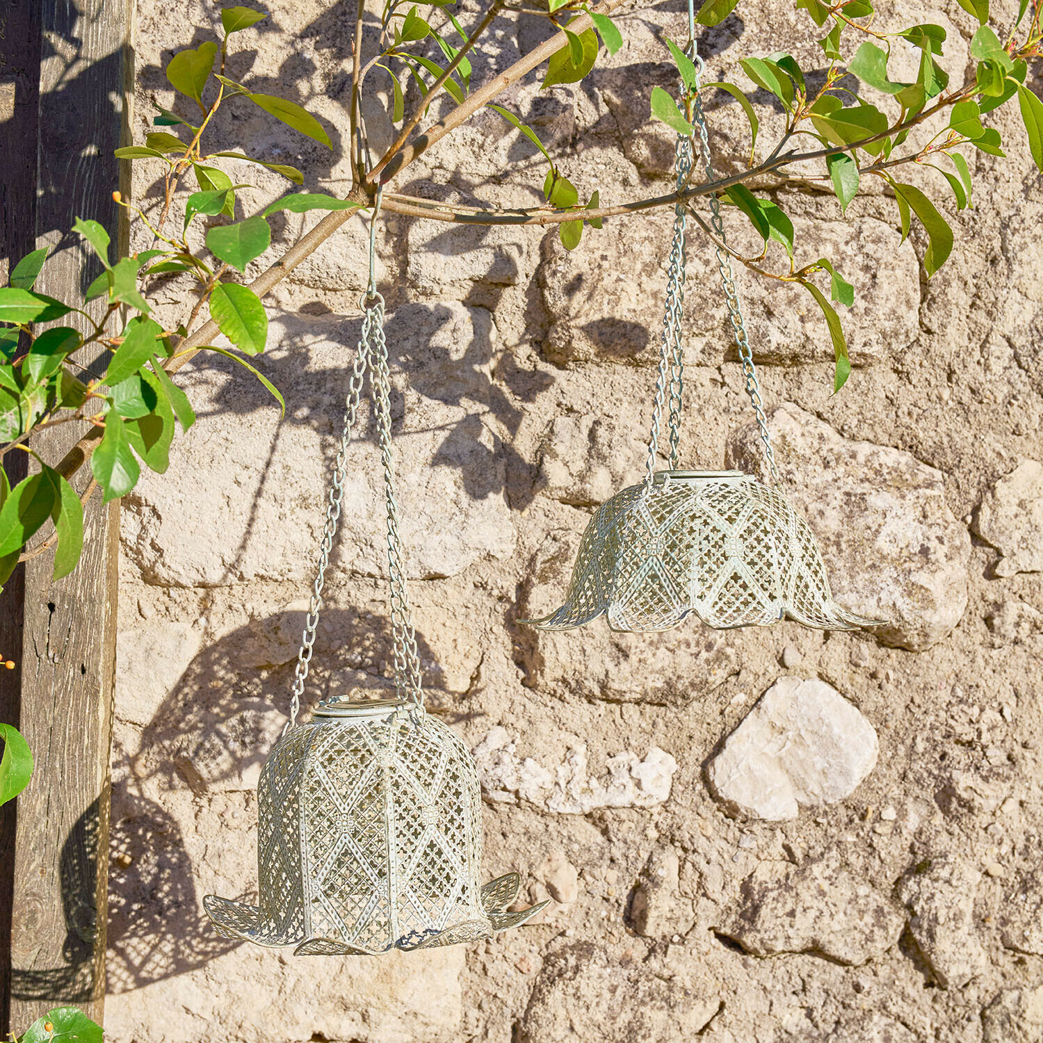 Twee hangende metalen lantaarns in klokvorm lichtgrijs tegen een stenen muur