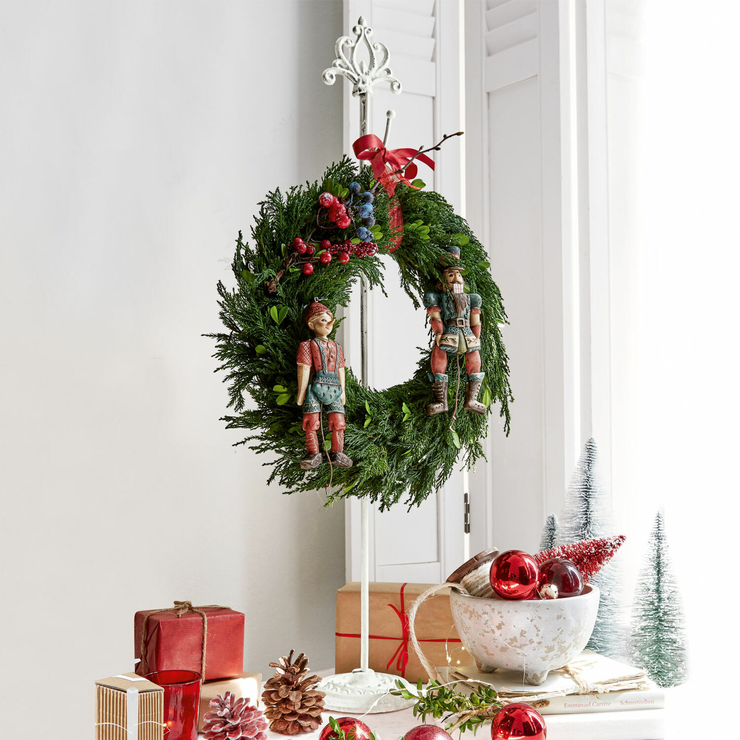 Kerstkrans van groene takken met rode en blauwe bessen en houten figuren op een witte standaard