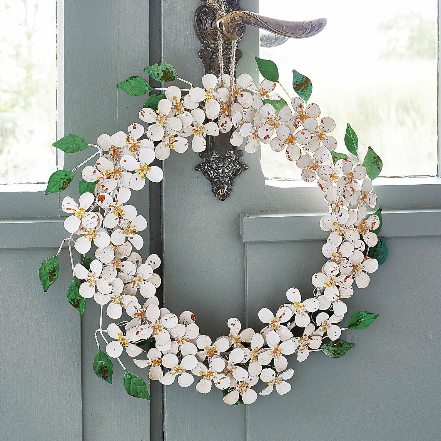 Krans Loanne Decoratieve metalen krans met witte bloemen en groene bladeren aan een grijze deur