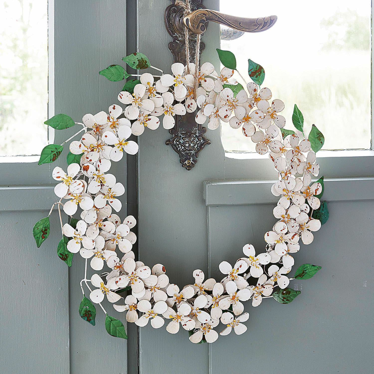 Krans Loanne Decoratieve metalen krans met witte bloemen en groene bladeren aan een grijze deur