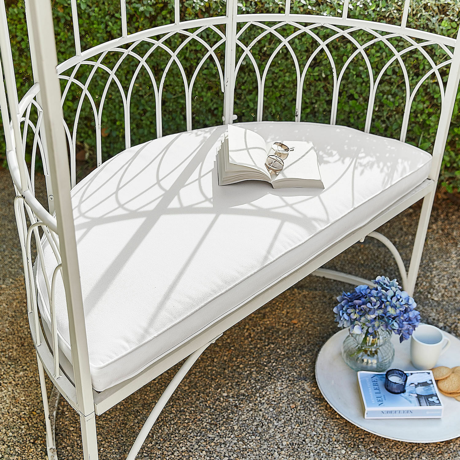 Witte metalen tuinbank met wit kussen, open boek, blauwe bloemen en mok op terras