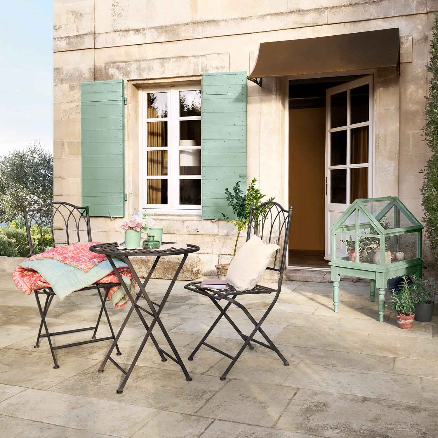 Metalen tuinset met tafel en stoelen op terras, groene mini-kas en pastelkleurige accessoires