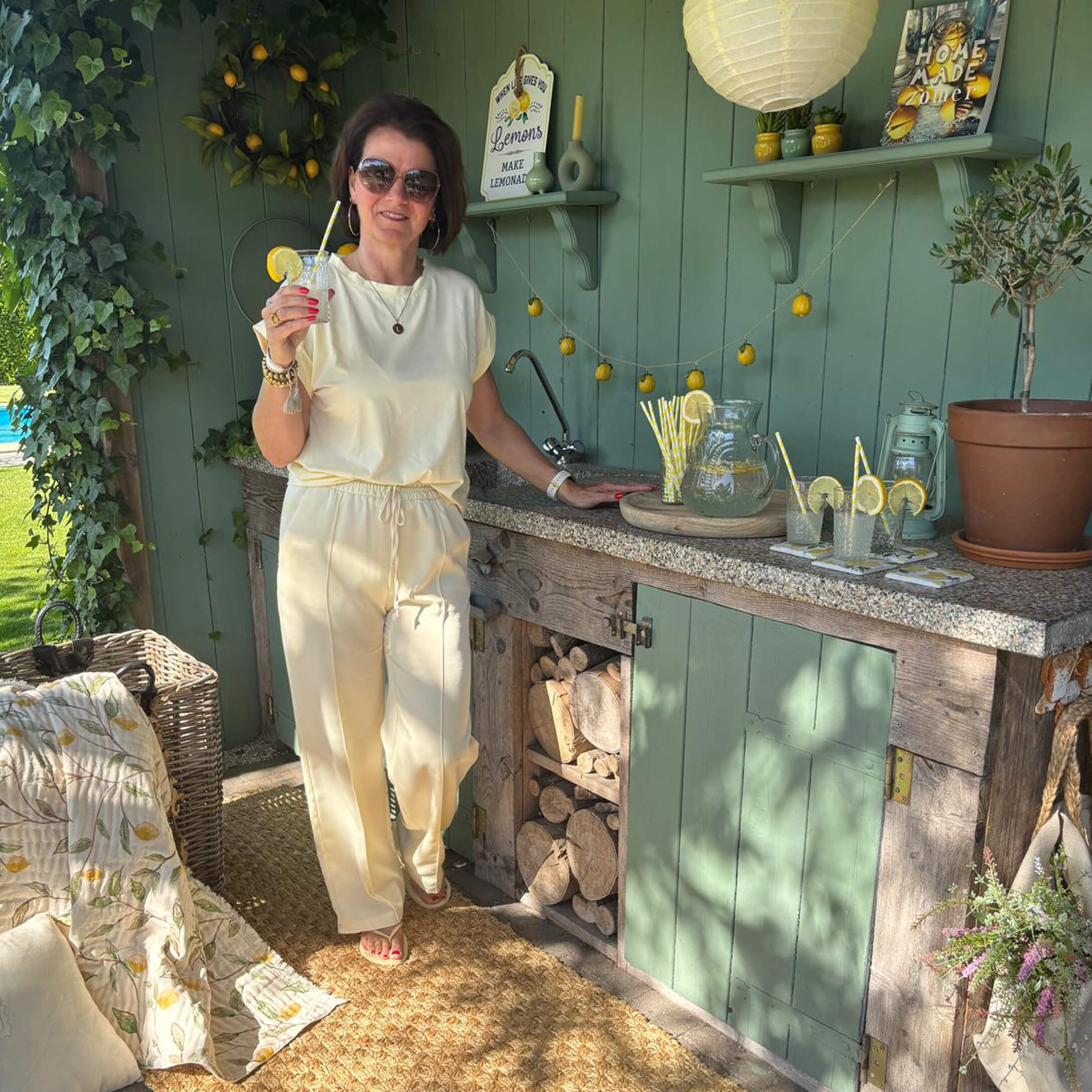 Vrouw in crème outfit bij groene buitenkeuken met limonade en citroendecoratie