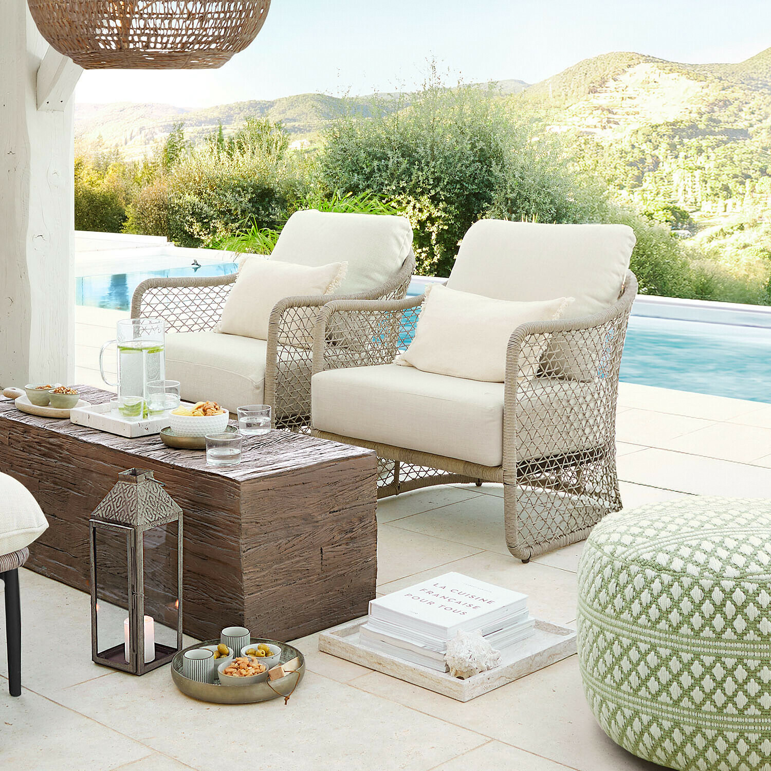 Twee beige stoelen van gevlochten materiaal op terras bij zwembad met houten tafel en groene poef