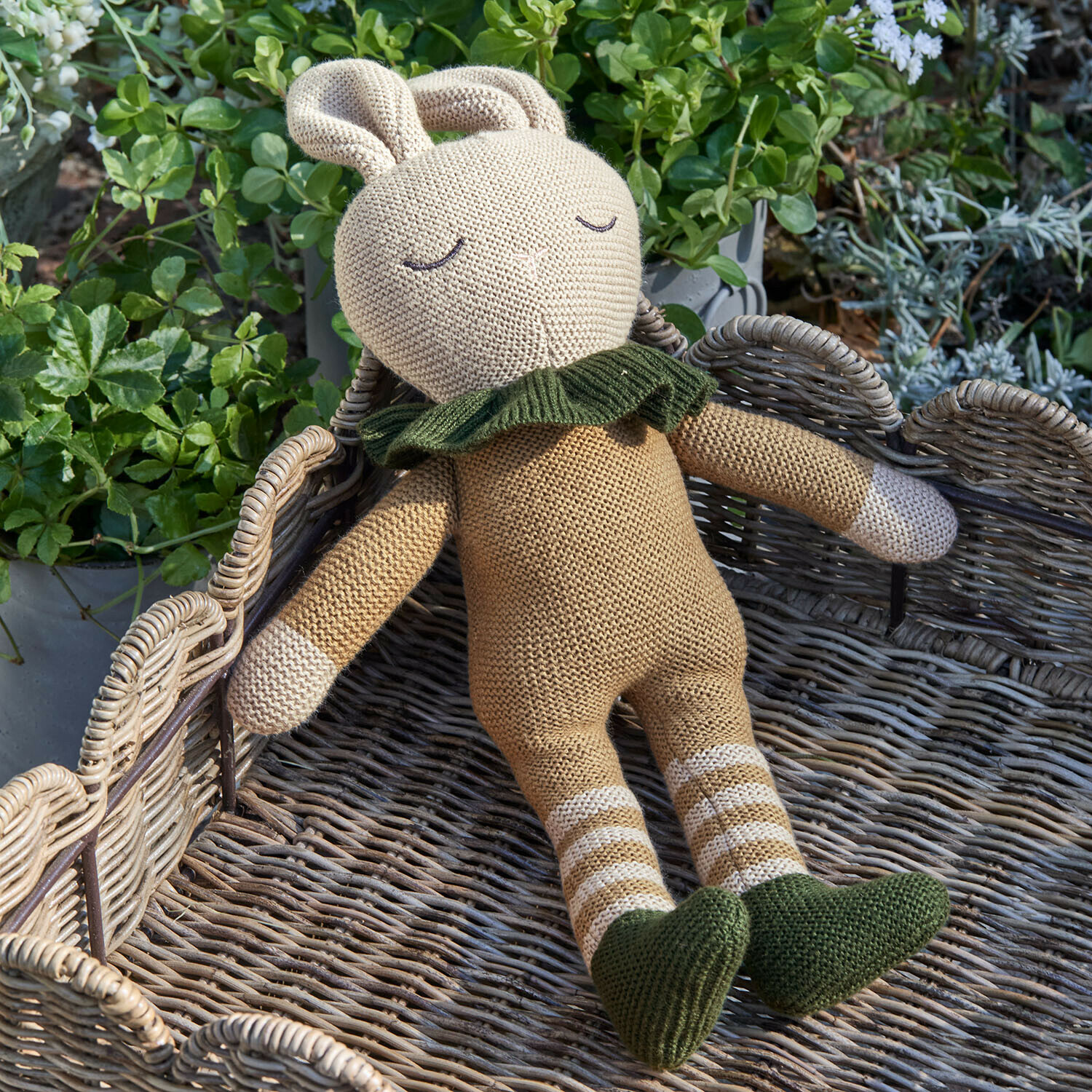Beige gebreide konijnenknuffel met groene schoenen en kraag op een rieten mand met planten eromheen