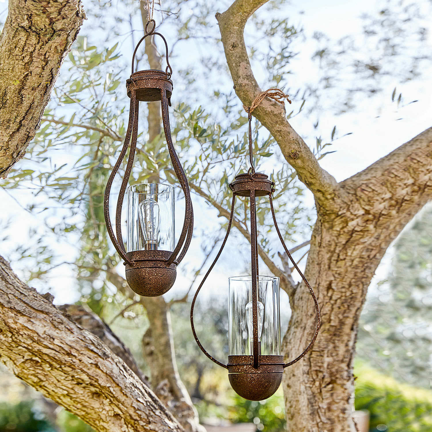 Twee hangende lantaarns van roestig metaal met helder glas in een buiten tuin