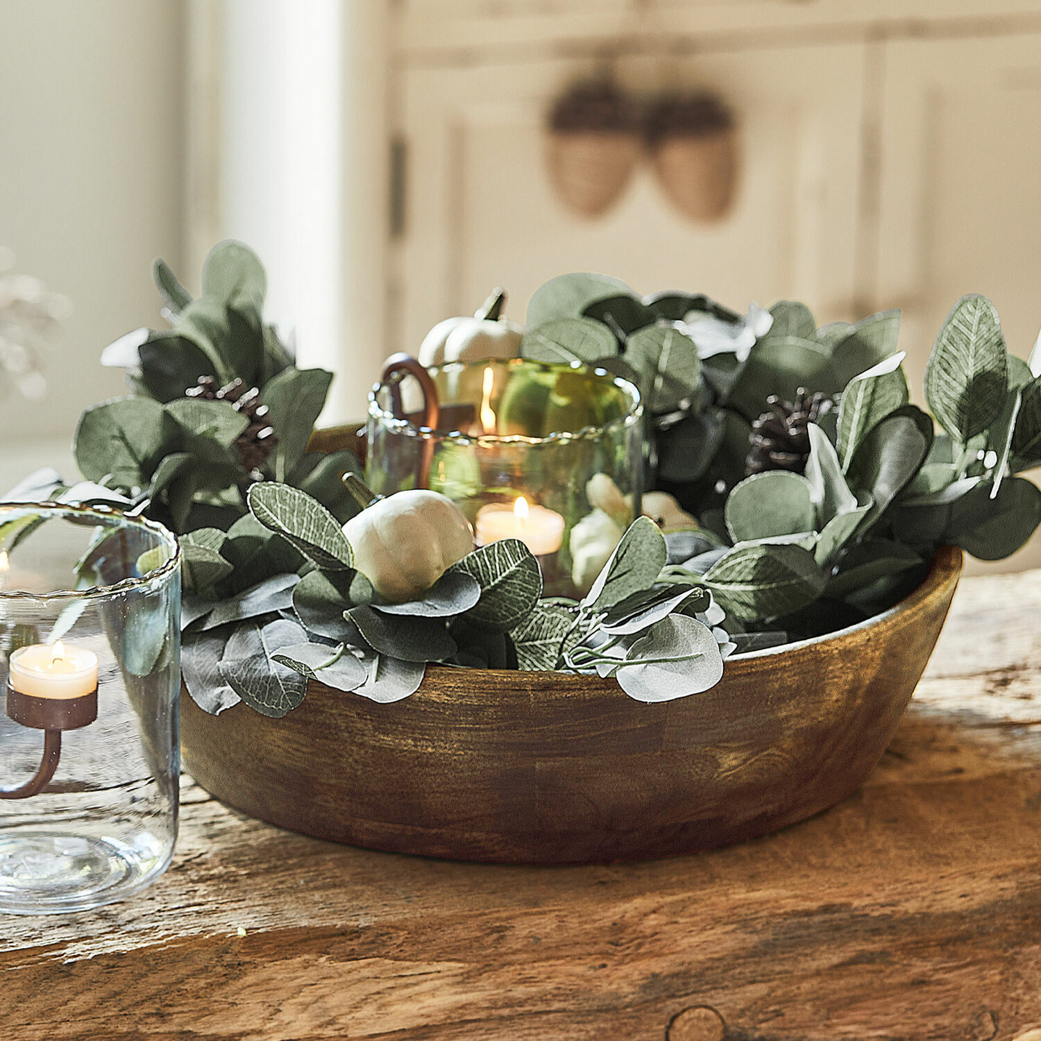 Decoratieve houten schaal met groene bladeren en kaarsen in glazen houders op tafel