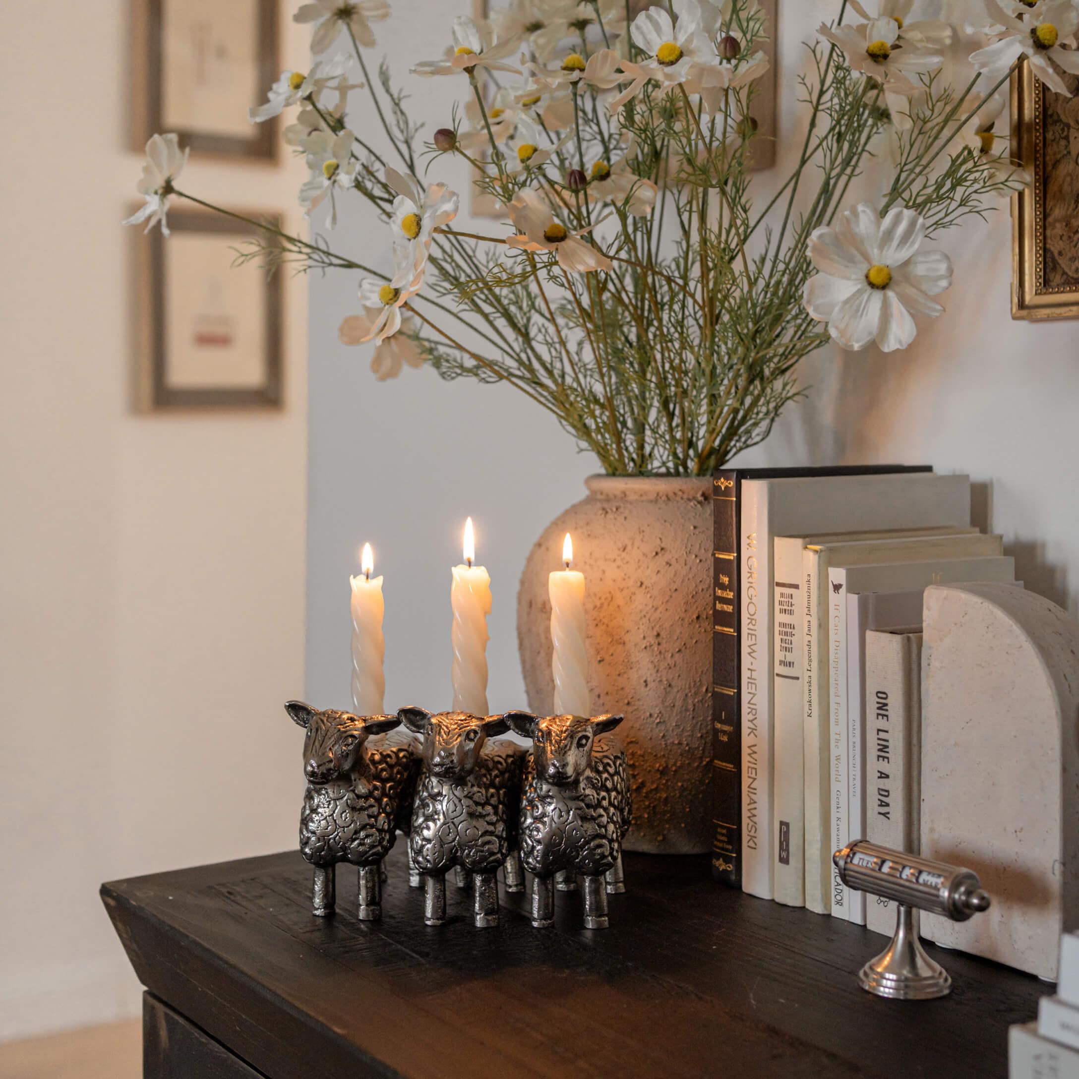 Metalen kandelaar in de vorm van drie schapen met witte kaarsen op een donkere houten tafel naast boeken
