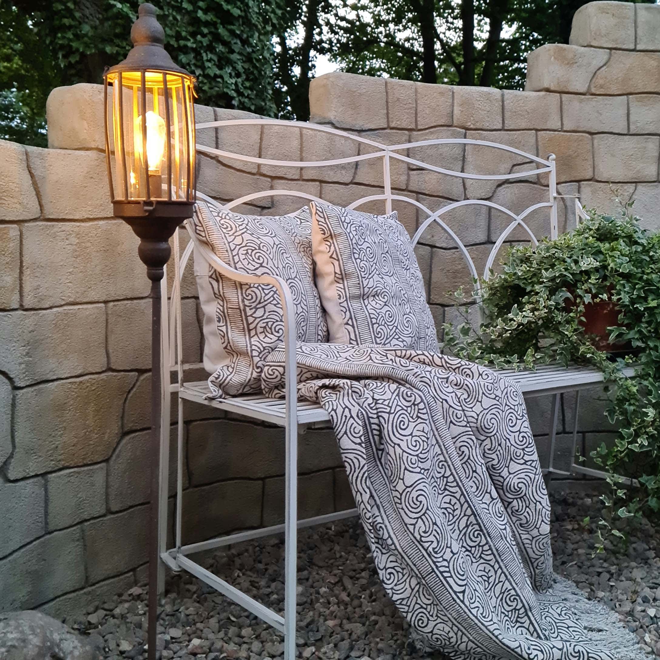 Witte metalen tuinbank met kussens en zwart-witte deken, verlichte staande lamp ernaast