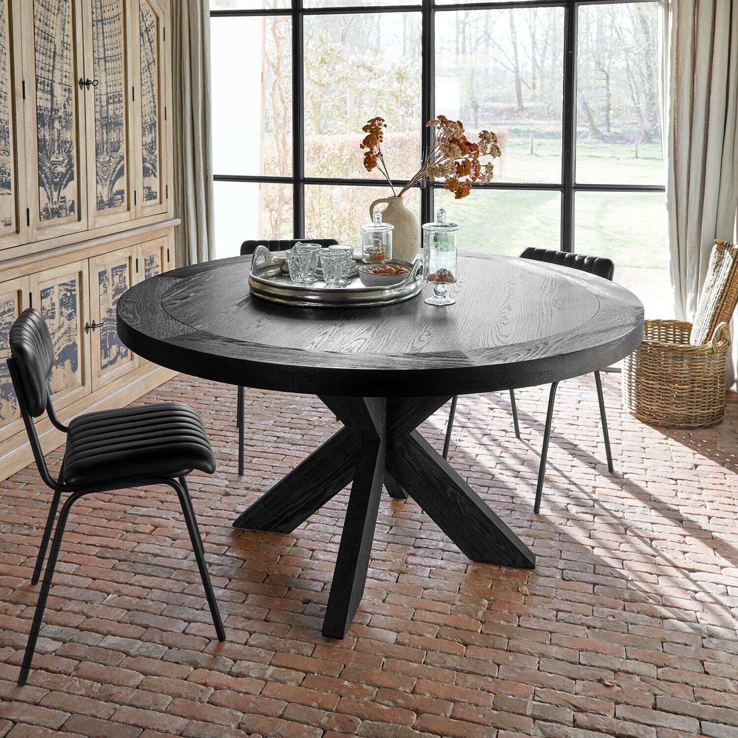 Tafel Darvoux Ronde zwarte houten tafel met decoratief dienblad en gedroogde bloemen in lichte kamer met bakstenen vloer