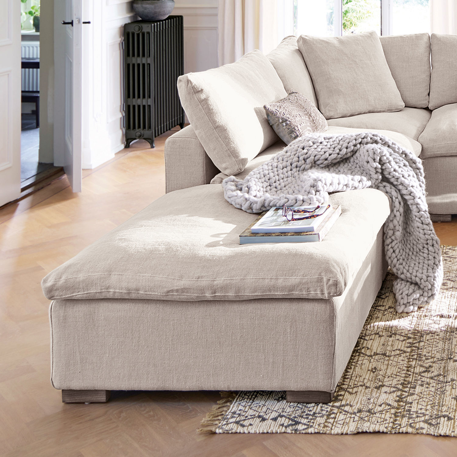 Beige stoffen hoekbank met zachte deken en kussens in lichte woonkamer met houten vloer