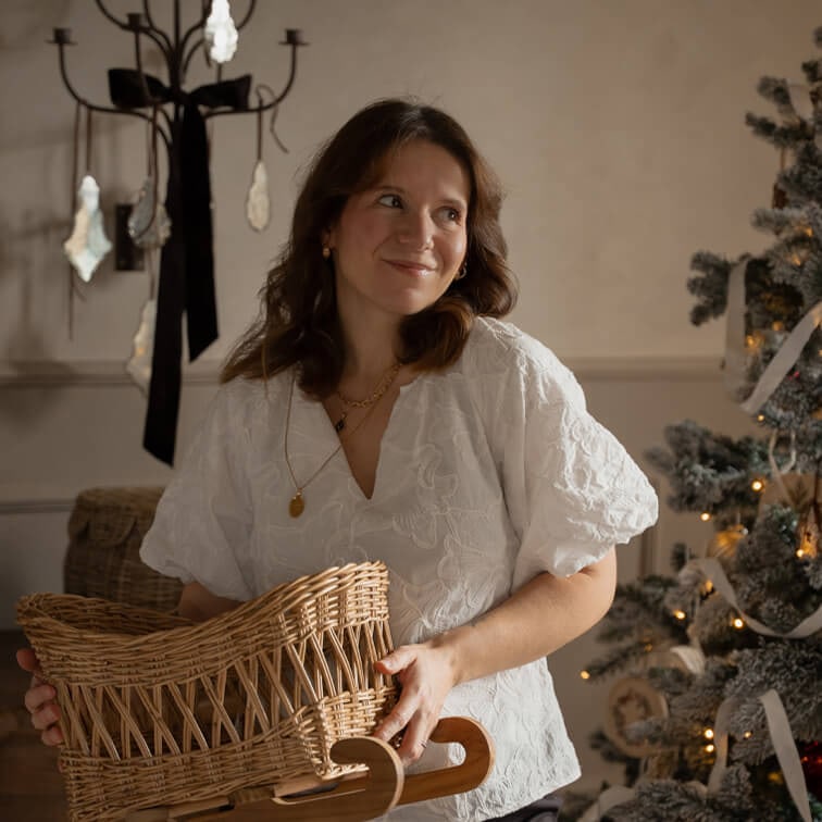 Vrouw in witte geborduurde blouse houdt rieten mand op houten slee bij versierde kerstboom