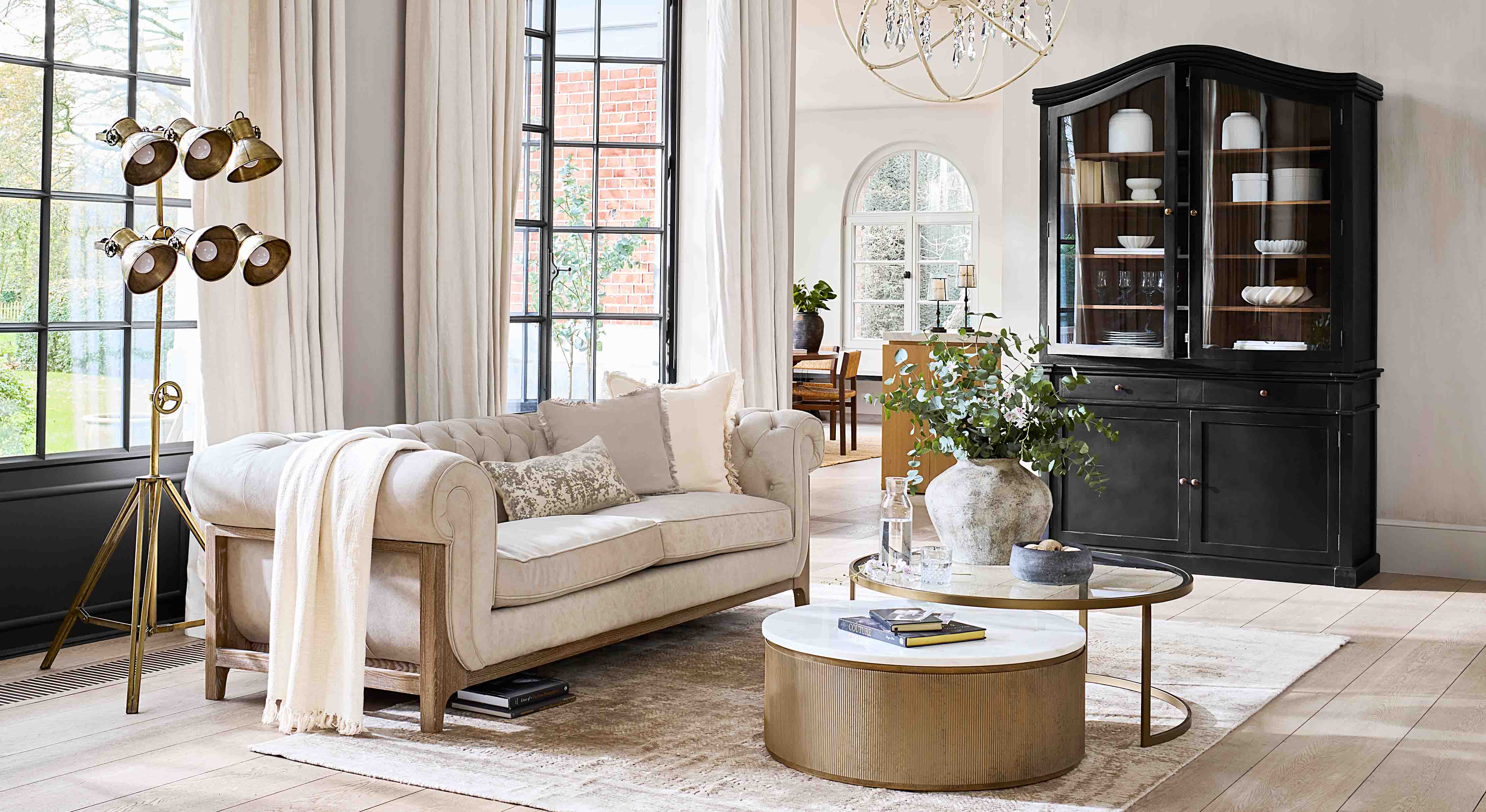 Beige bank met houten frame in lichte woonkamer met zwarte vitrinekast en gouden vloerlamp