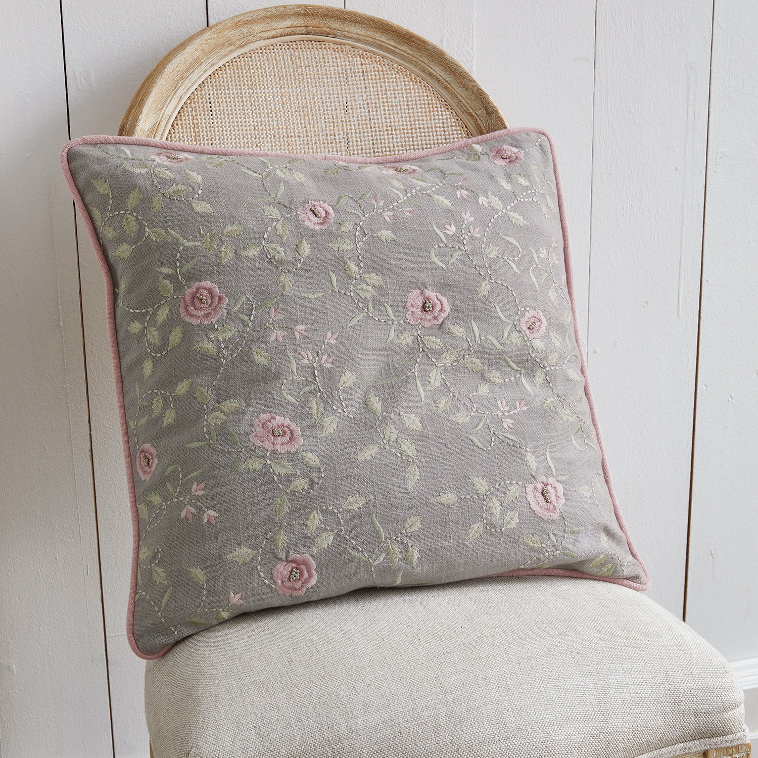 Grijs sierkussen met roze bloemenborduursel op beige stoel, woontextiel voor gezellige ruimtes