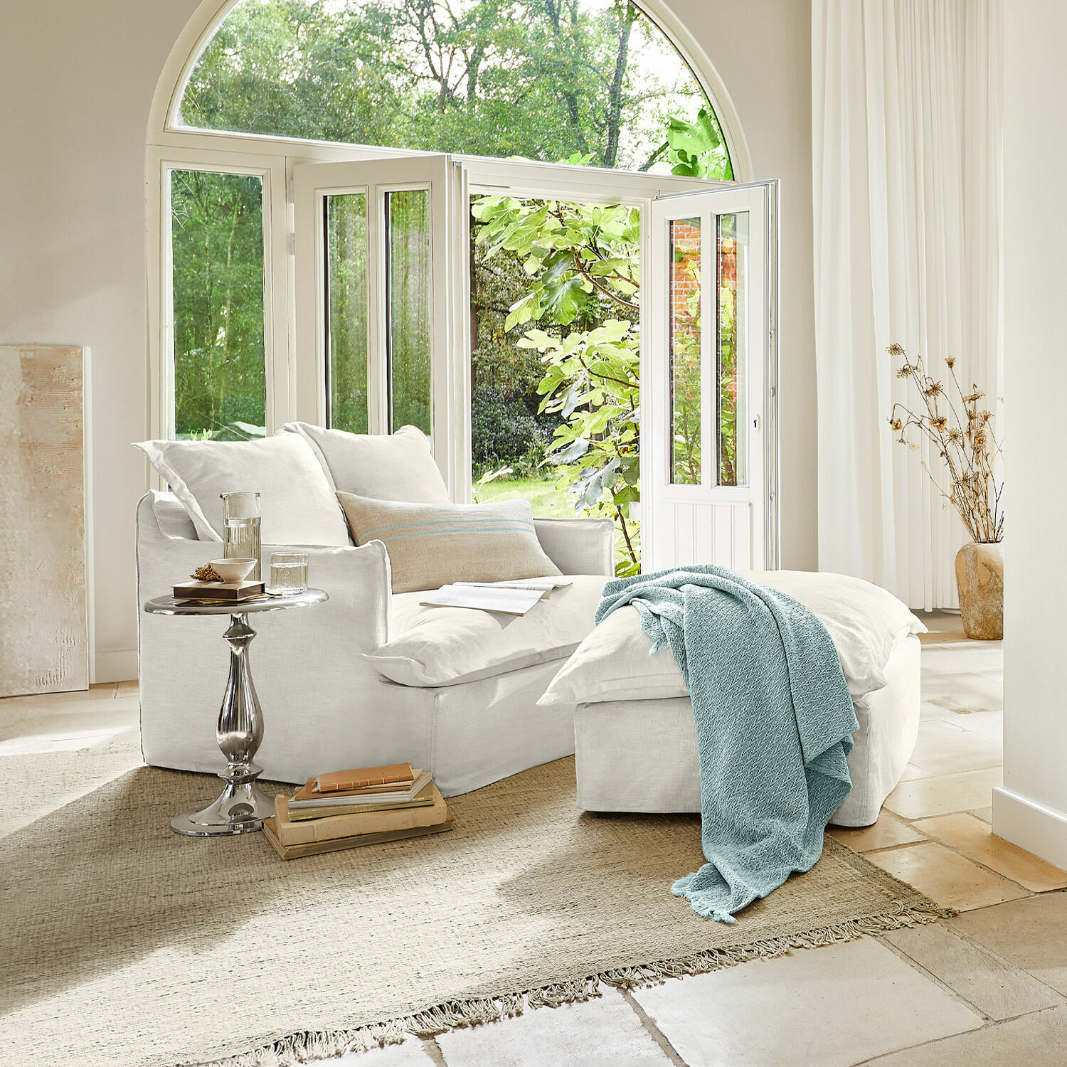 Fauteuil met hocker Avignon Witte katoenen fauteuil met voetenbank en blauwe deken in lichte woonkamer met groot raam